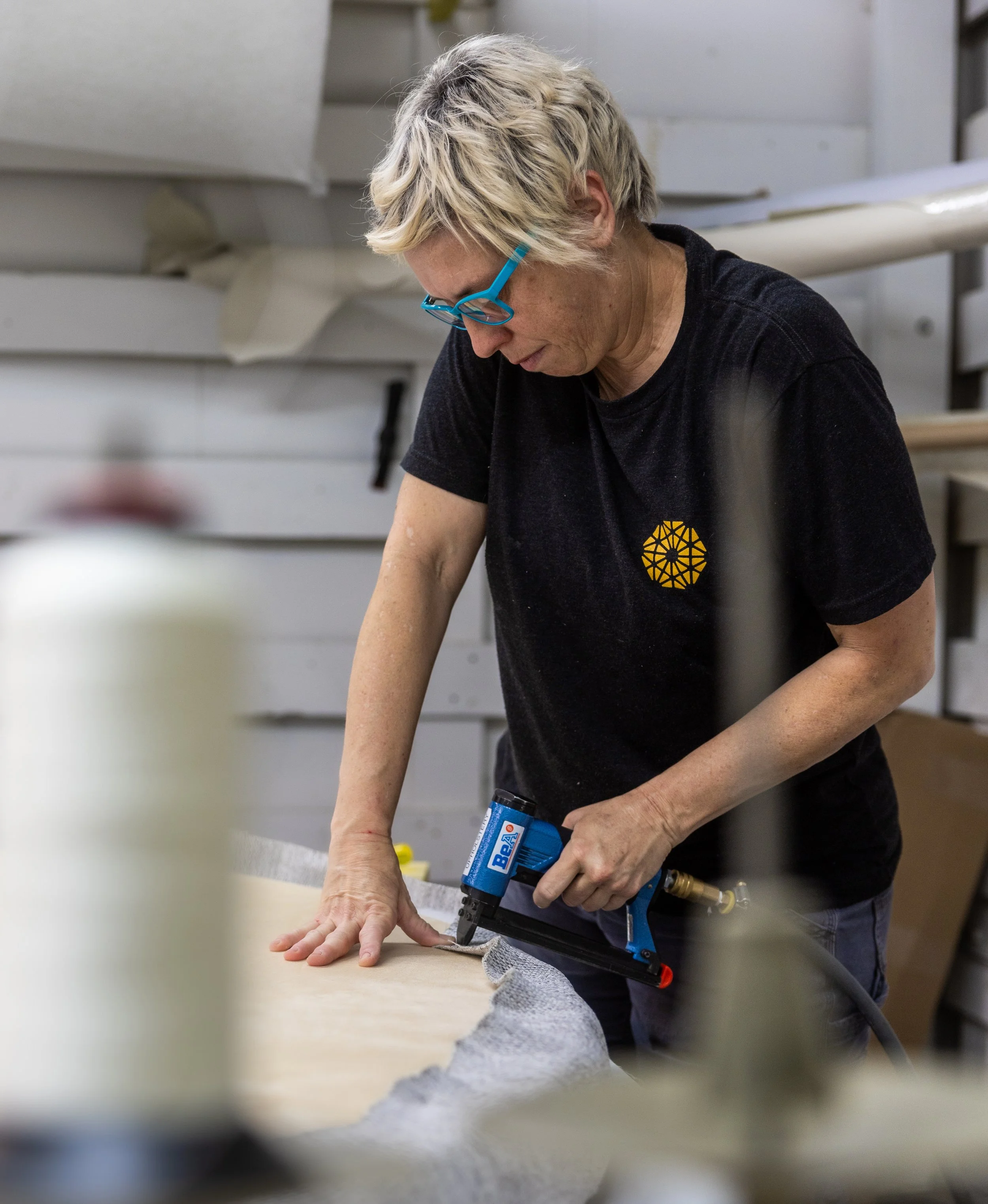 A woman with short blonde hair wearing a black Tecture t-shirt and bright blue glasses, uses a pneumatic stapler to secure fabric to a custom furniture piece, focusing on precision in a workshop setting.