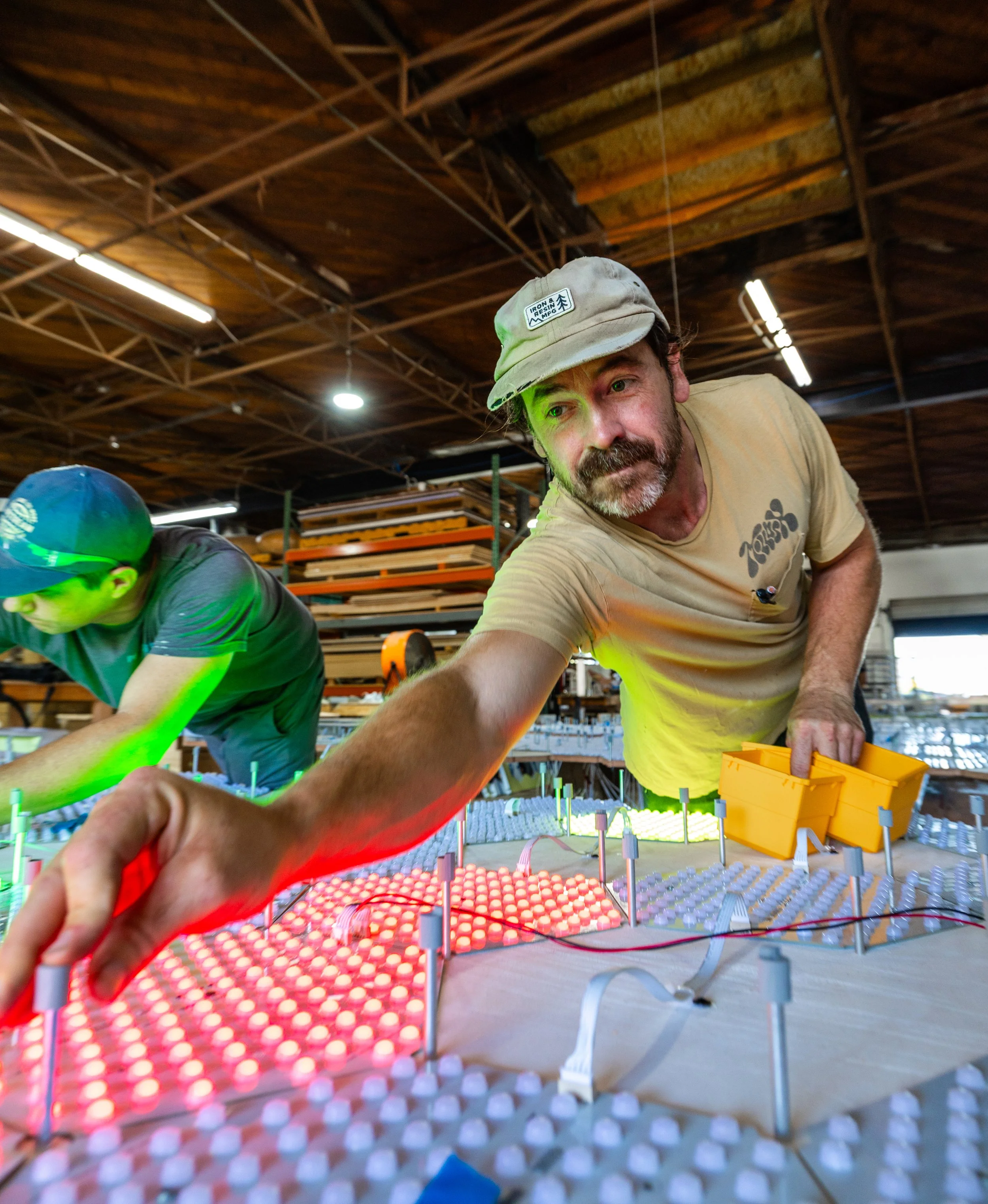 A man with a brown and gray mustache, wearing a pale yellow t-shirt and gray hat in a workshop install and adjust LED lighting components on a custom panel, working with colorful illuminated elements and wiring.