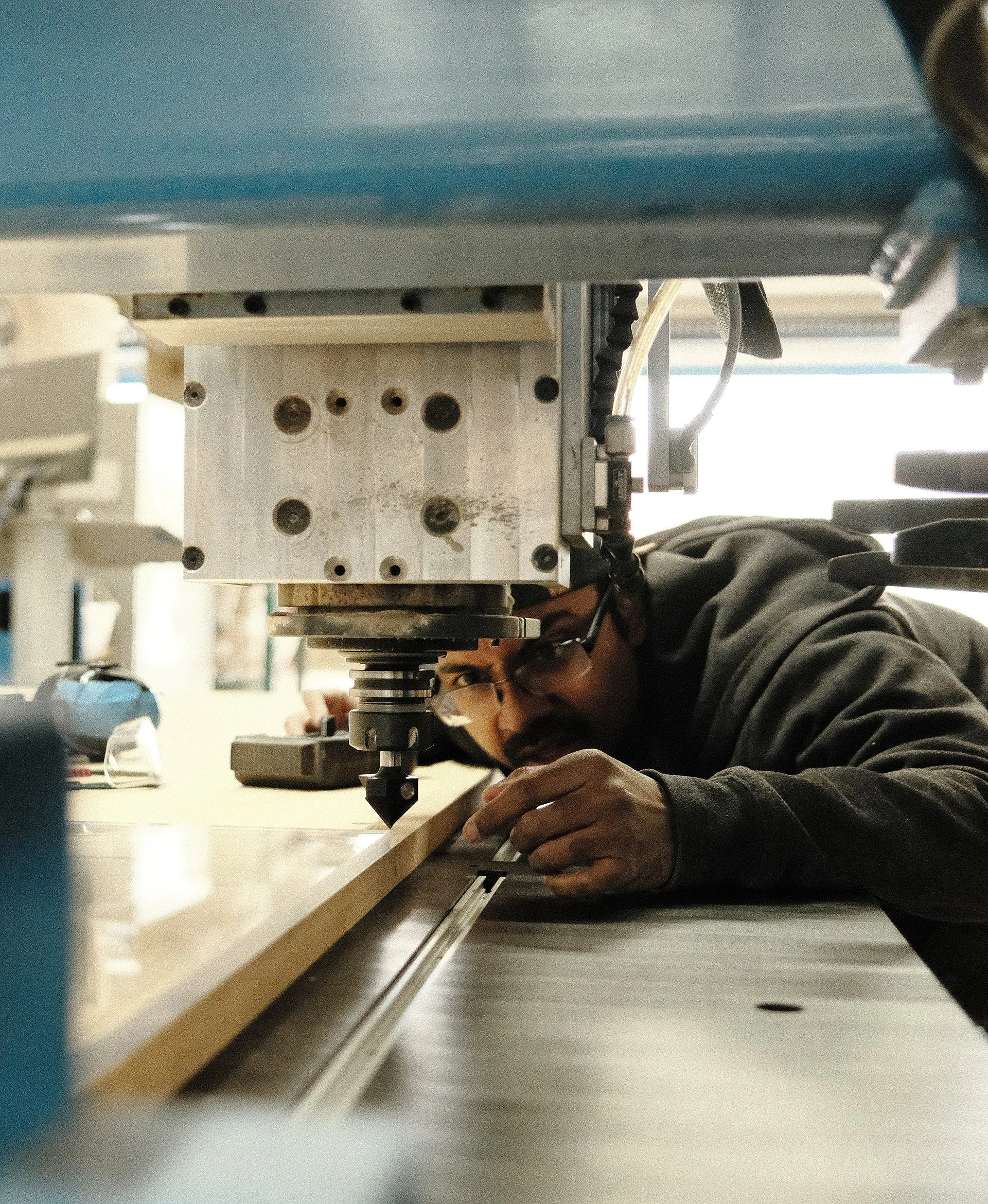 A man wearing a dark gray sweatshirt and glasses, aligns material beneath a CNC machine, carefully setting up a precision cut in a workshop.