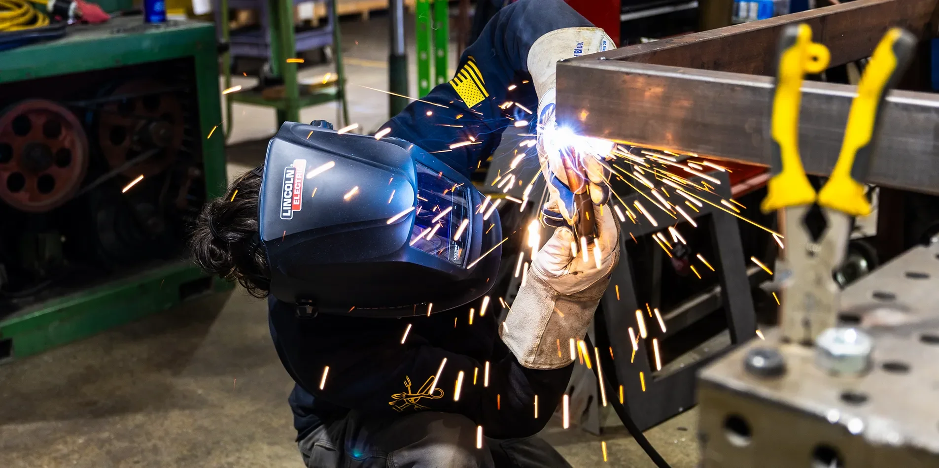 Fabricator welding a structural steel assembly in the shop with sparks flying and protective head and face gear
