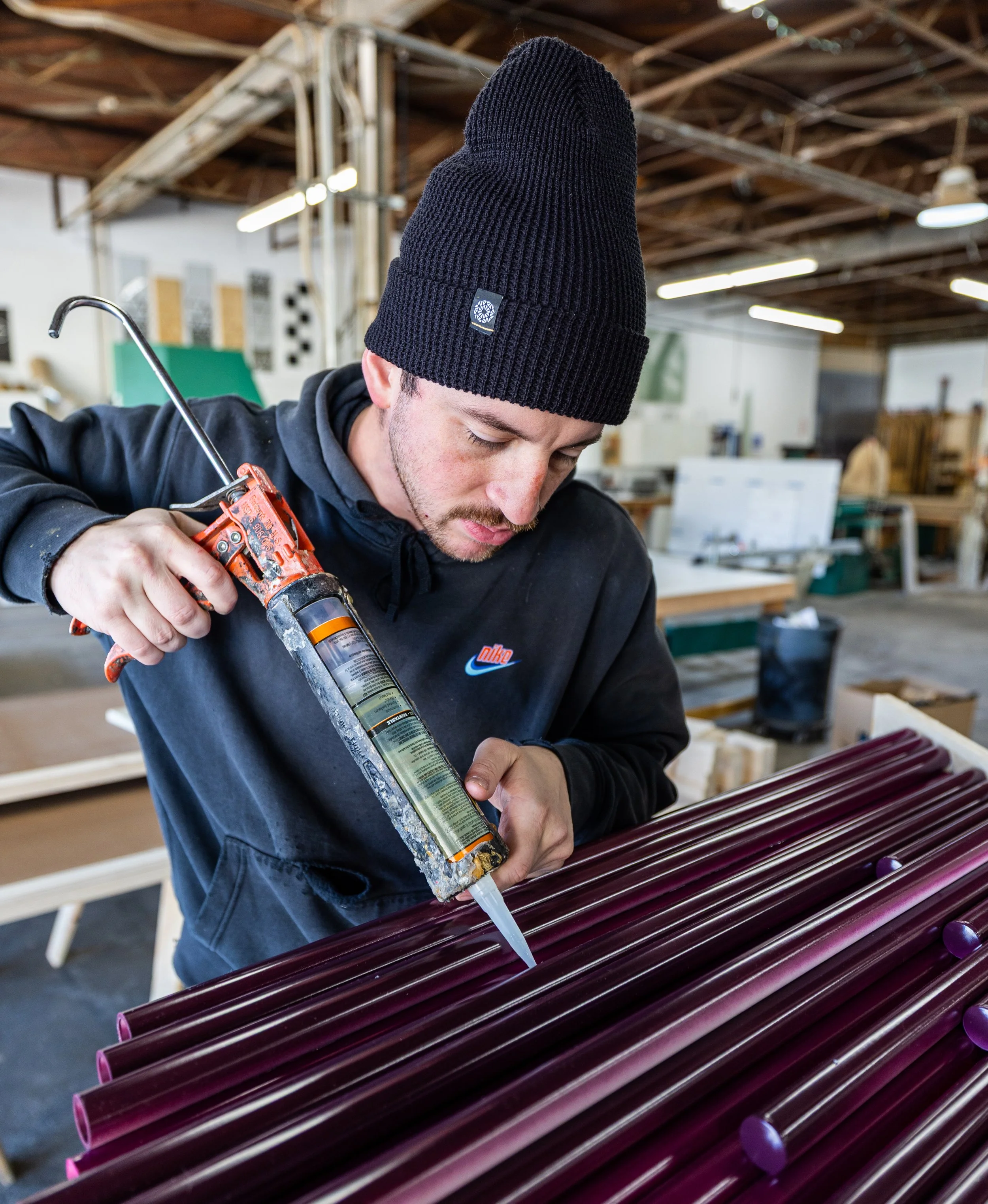 A man with a mustache wearing a black beanie and black sweatshirt, applies adhesive to colored acrylic rods in a workshop, assembling components with precision and care.