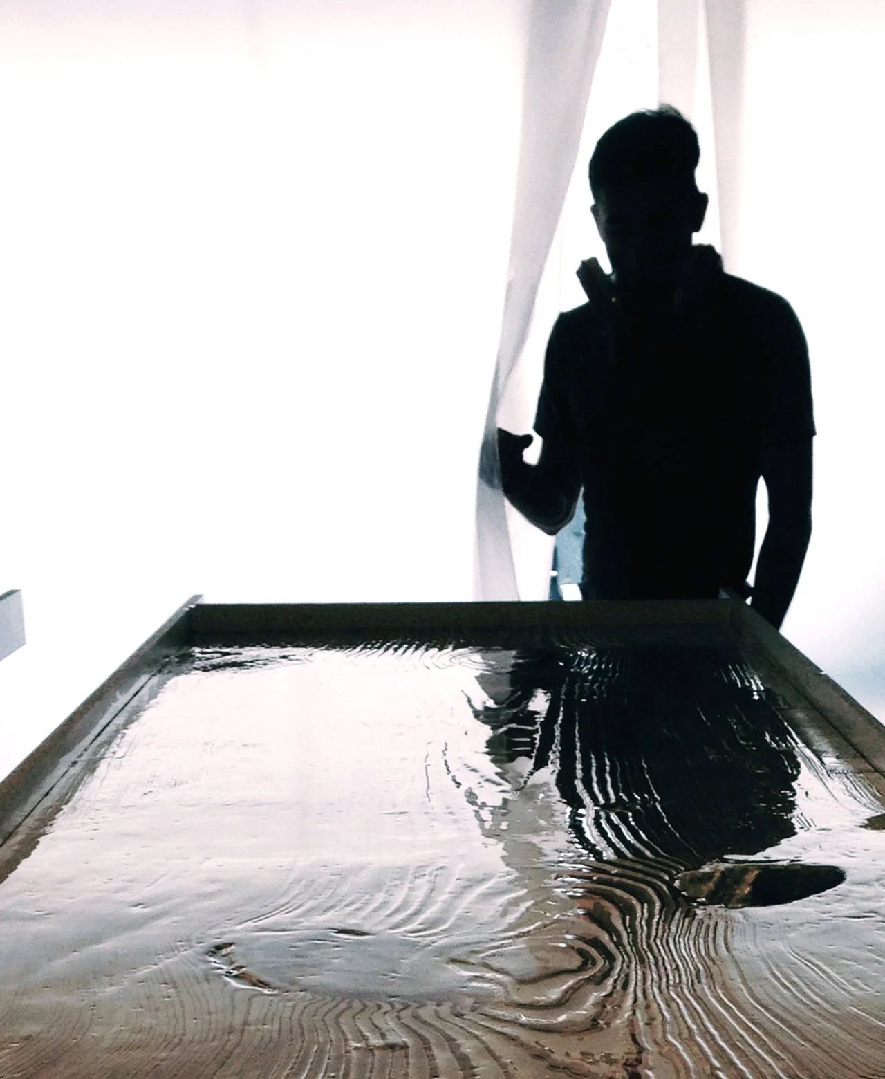 A man in silhouette working on wooden table with metal frame pouring resin. 