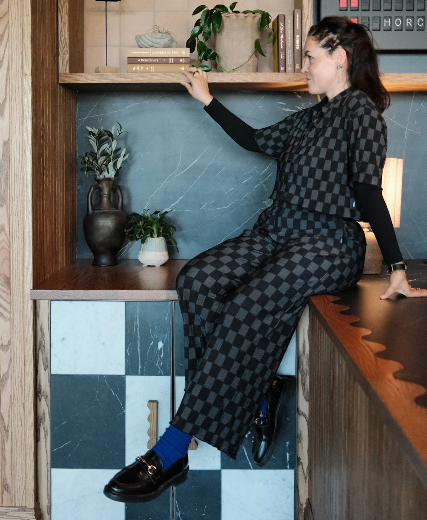 A woman with dark brown hair, wearing a black and gray checkered outfit and blue socks sits on a custom built-in wood counter with shelving, arranging books and decor in a modern interior with mixed materials and greenery.
