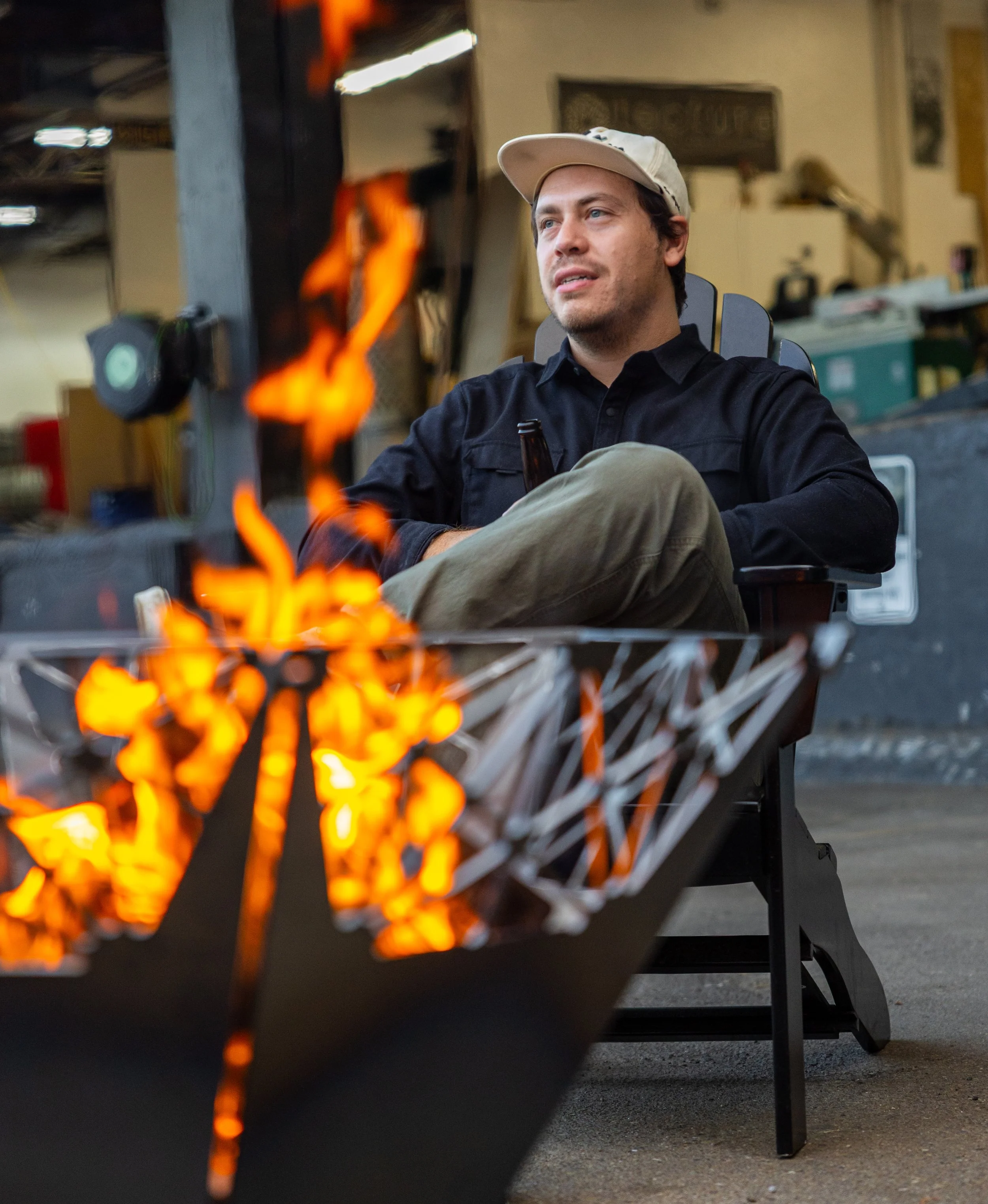 A man smiling with a light beige trucker hat, black long sleeve shirt and olive green pants relaxes in a chair behind a metal fire pit with a geometric pattern cut out, with visible flames, showcasing custom fabrication work in an industrial setting.