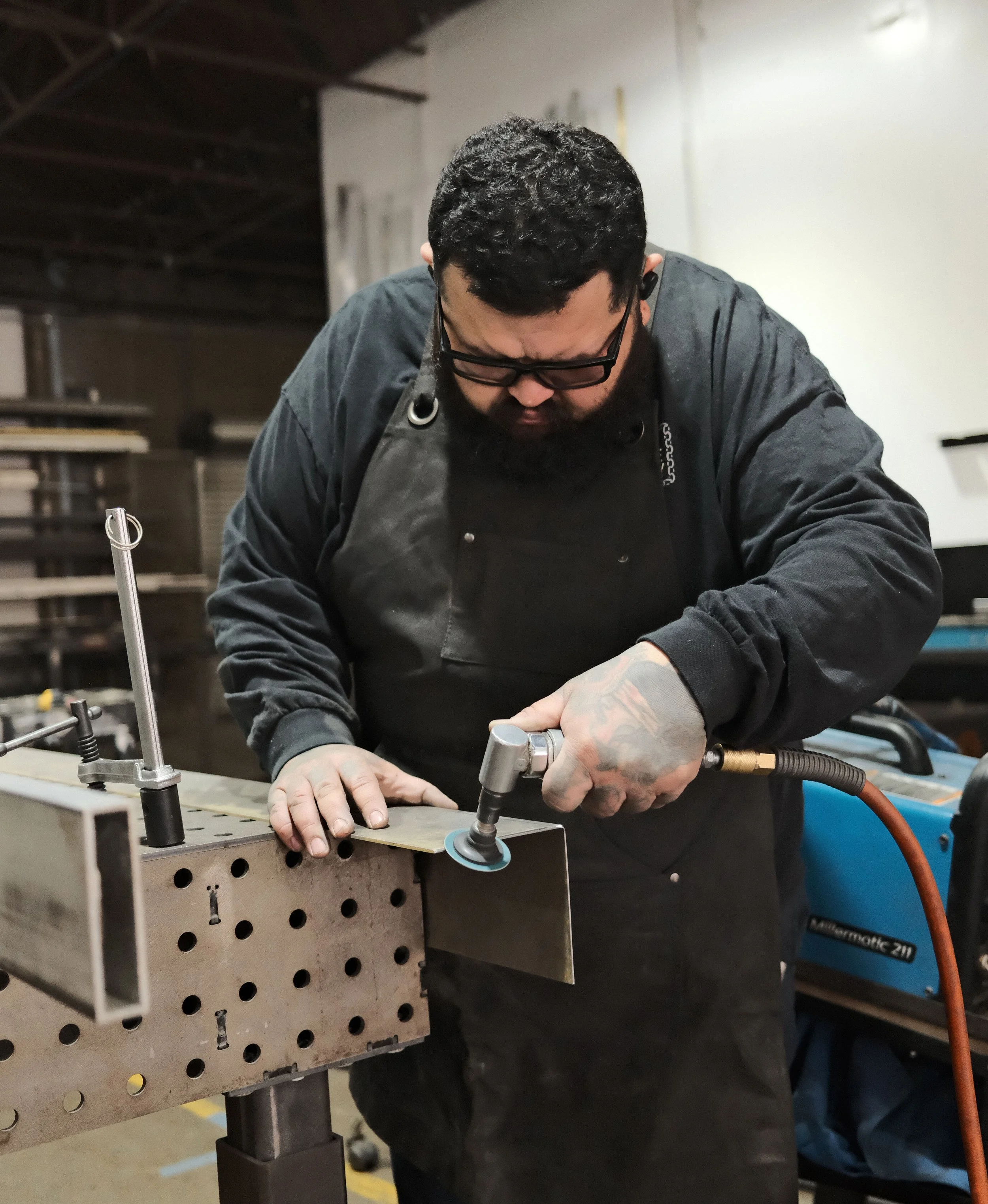 A man with short black hair wearing a black long sleeve shirt and welding vest, in a workshop uses a handheld grinder to finish a steel component, focused on precision and craftsmanship.