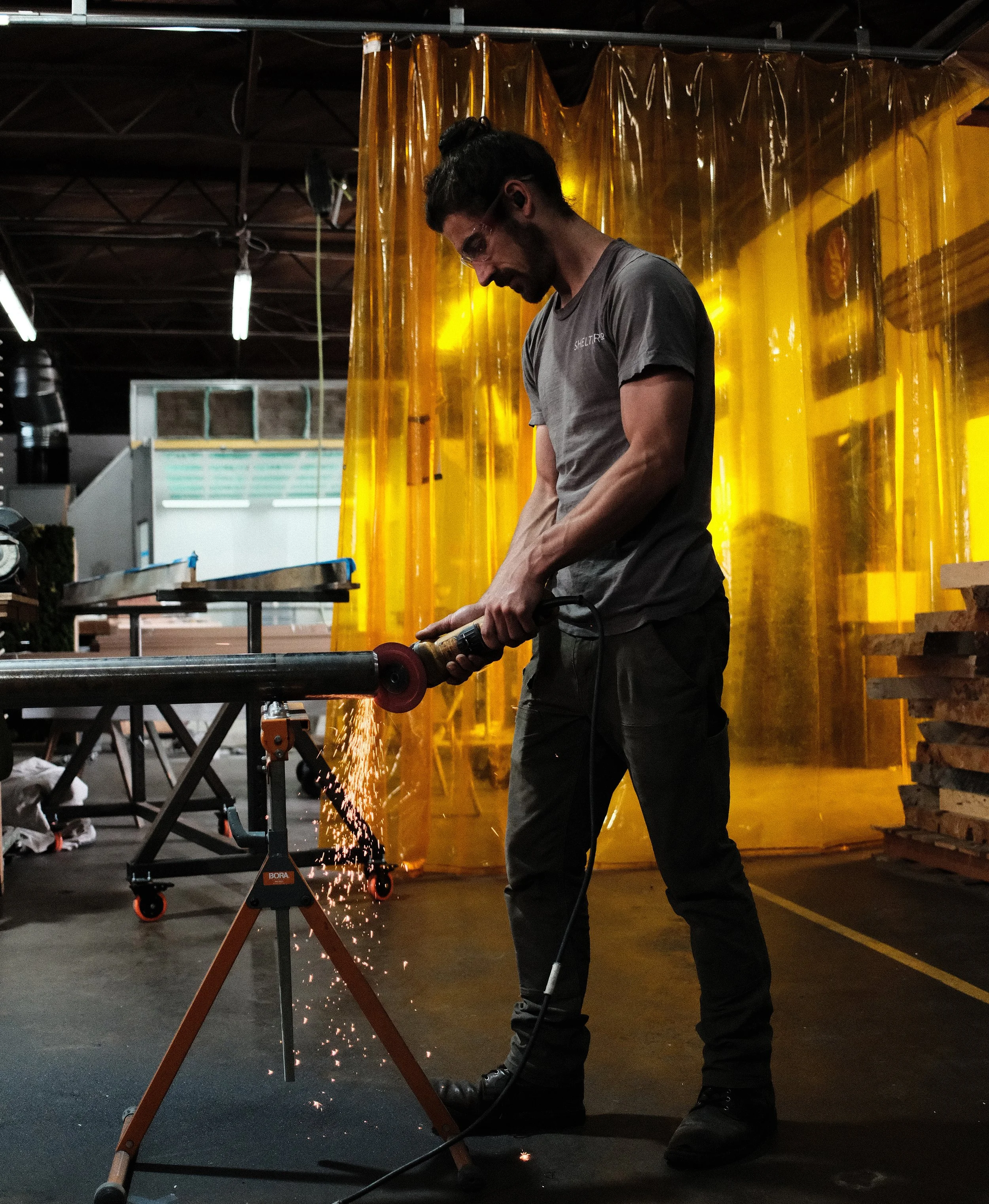 A man with dark long hair in a bun, wearing a short sleeve gray t-shirt, uses an angle grinder on a steel pipe in a workshop, creating sparks against a backdrop of industrial equipment and yellow welding curtains.