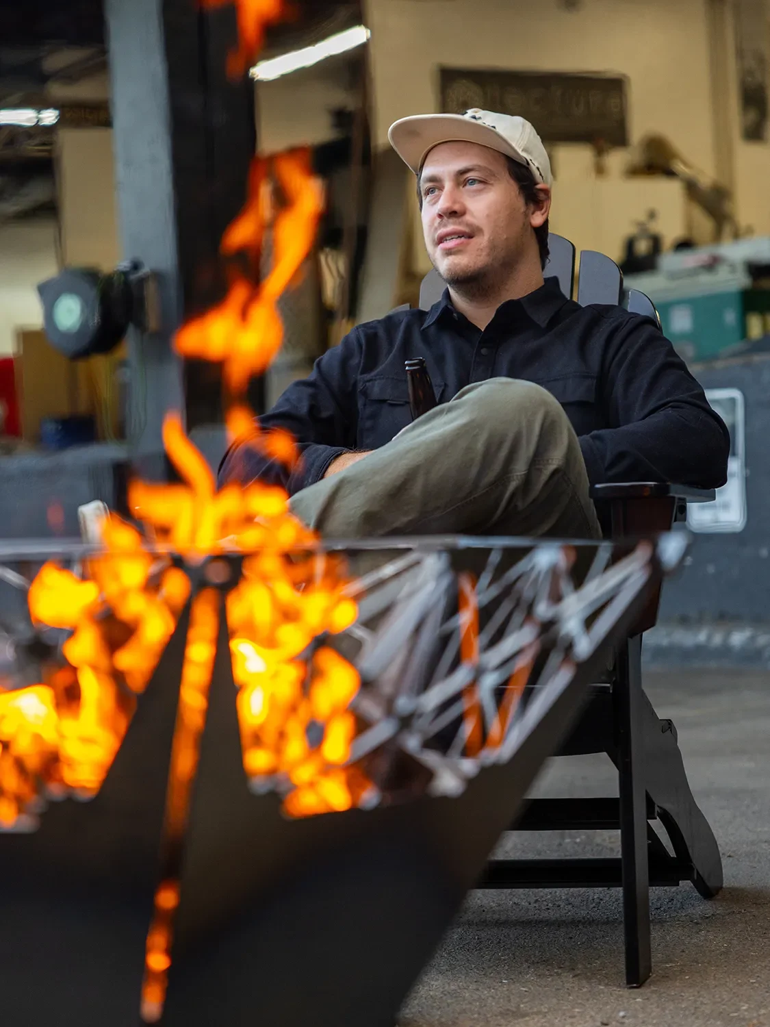 Alex Resler seated beside a lit custom steel fire pit with geometric cutouts at the Tecture shop