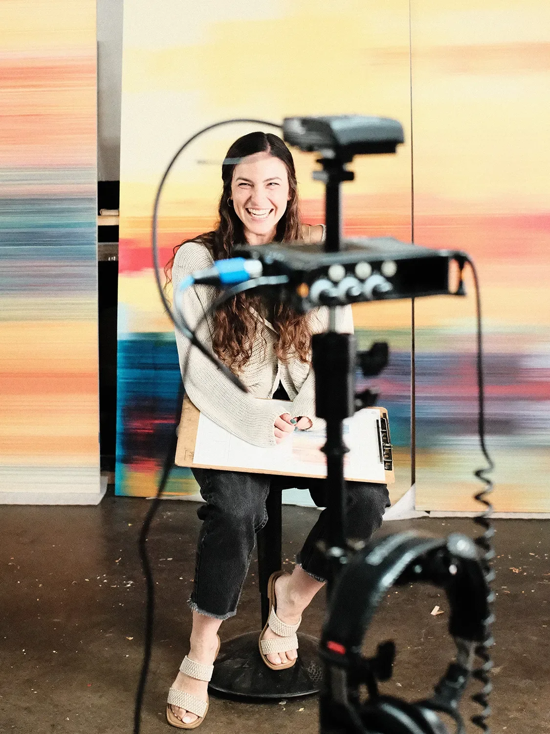 Jenna Matus laughing during filming, seated in front of colorful abstract artwork at Tecture