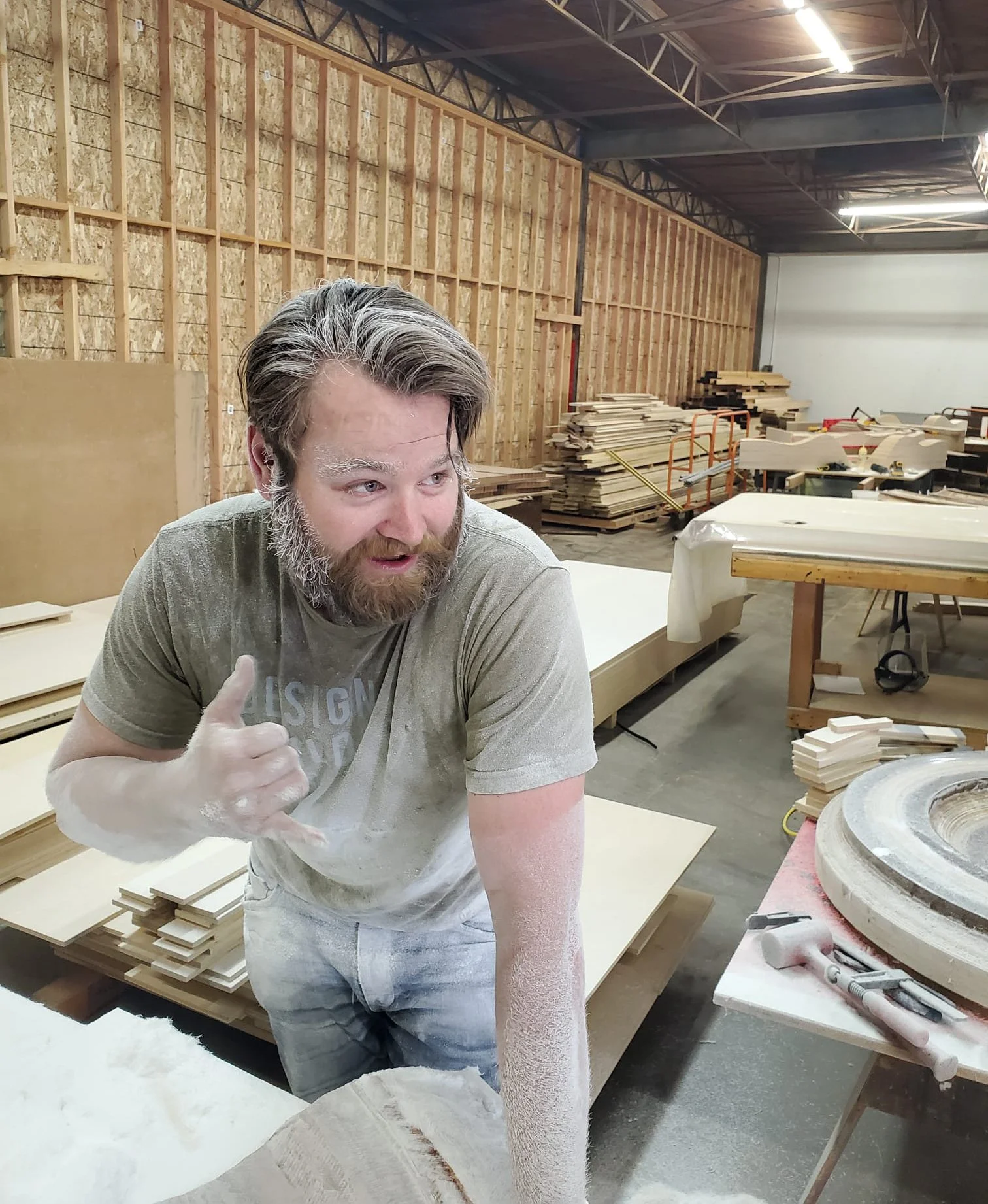 A man with a beard and mustache standing in a warehouse with stacks of wood behind him, wearing a grey T-shirt, holding up a shaka sign and is covered in white dust..