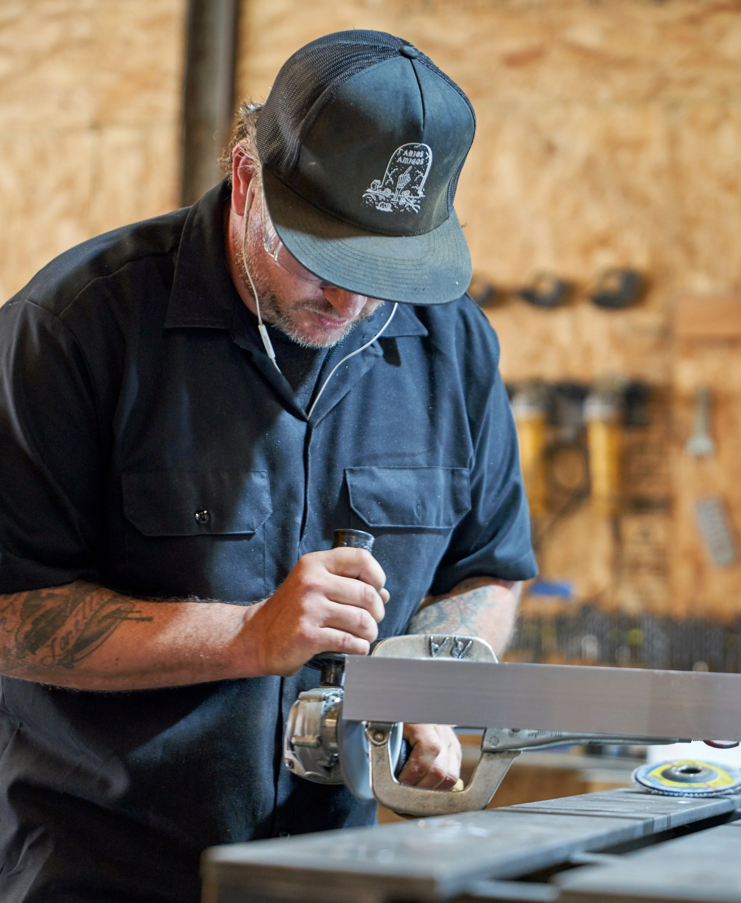 A man wearing a black button down t-shirt and black hat with tattoos on his arms, stands in a workshop using a handheld grinder to shape a metal piece, focusing on detail and precision craftsmanship.