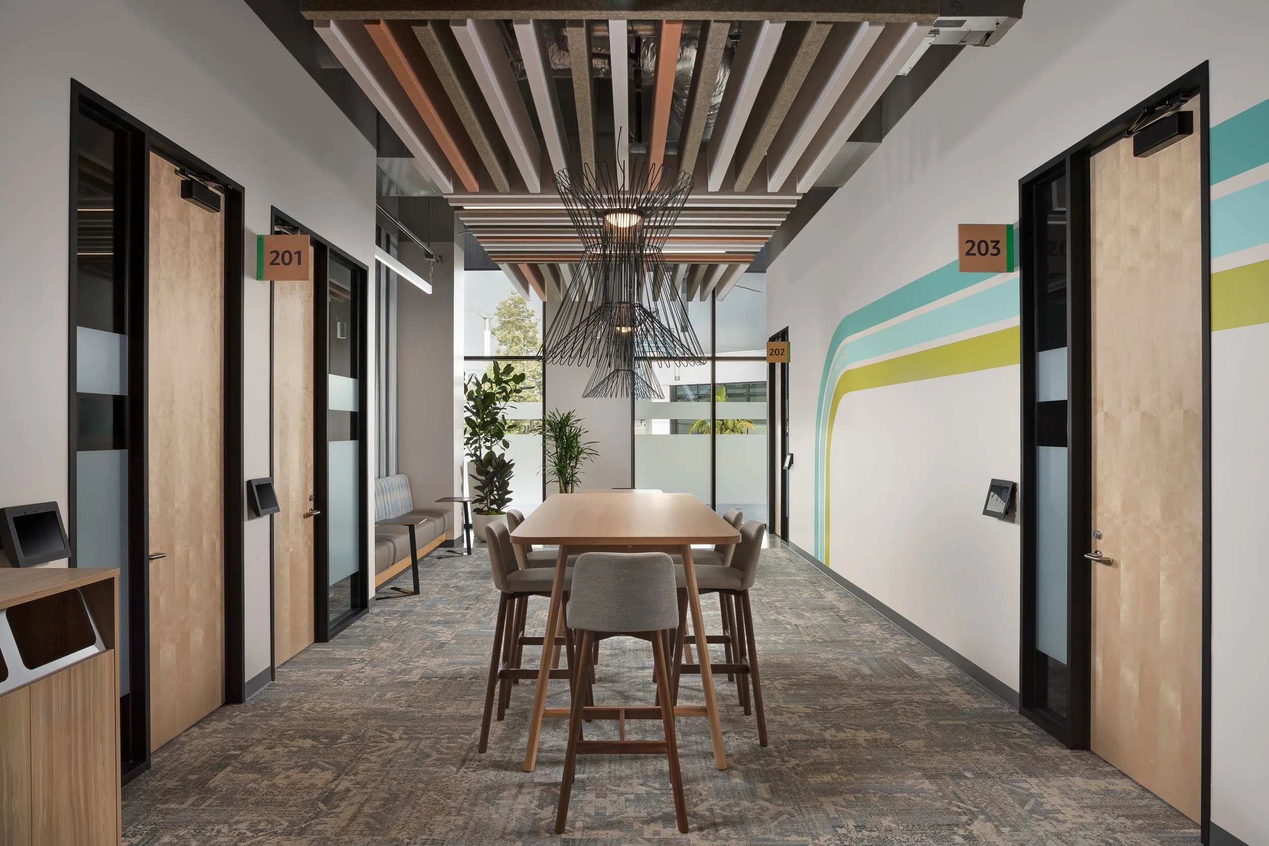 Office corridor with a wood communal work table, blue and green wall graphics, and a custom suspended ceiling feature.