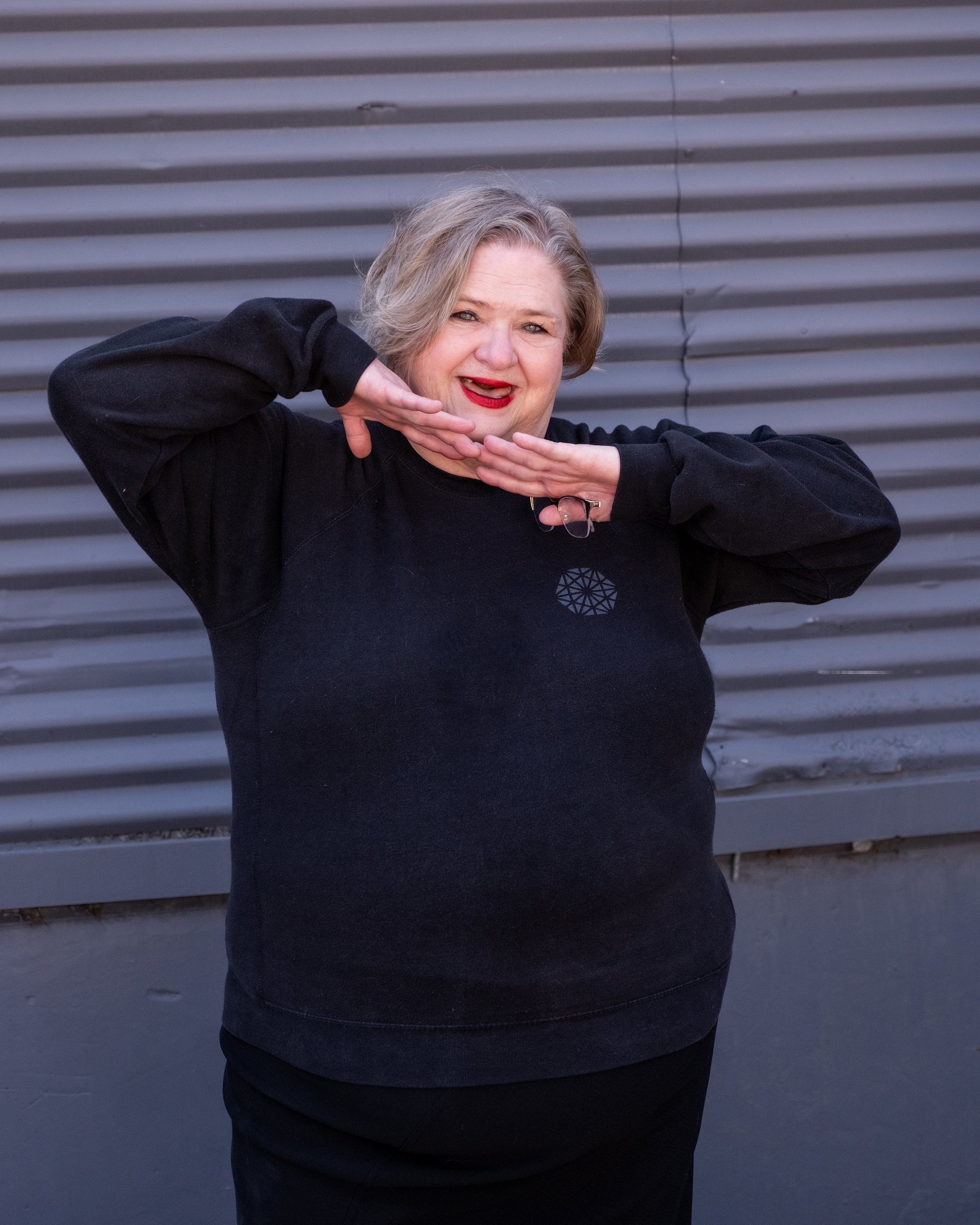 A woman with short gray hair smiling and making a playful gesture with her hands on her face, standing outdoors in front of a metallic wall.