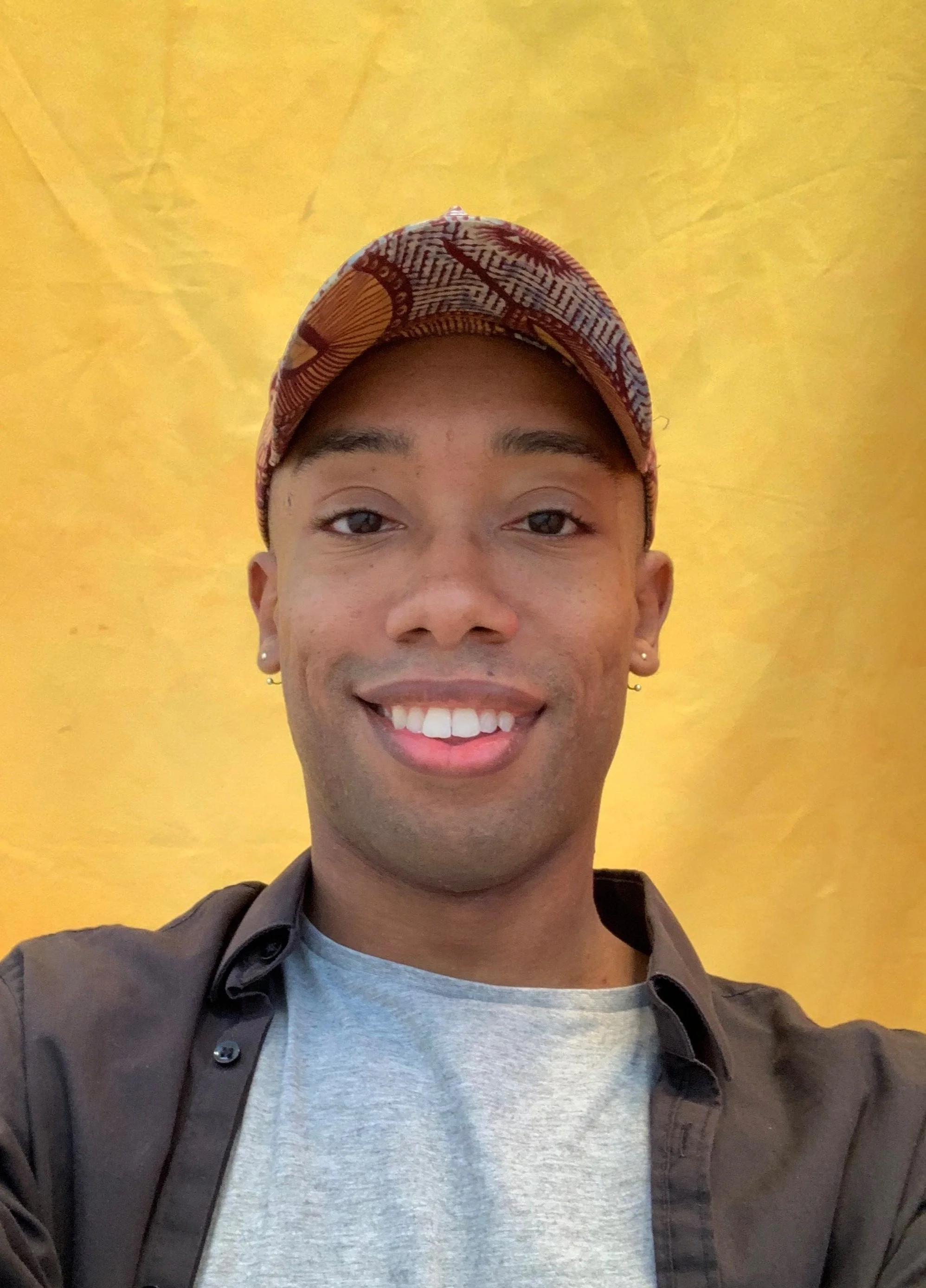Founder of Pheme AI, Gyasi Patterson, smiling while wearing a patterned cap and black shirt, against a yellow background.