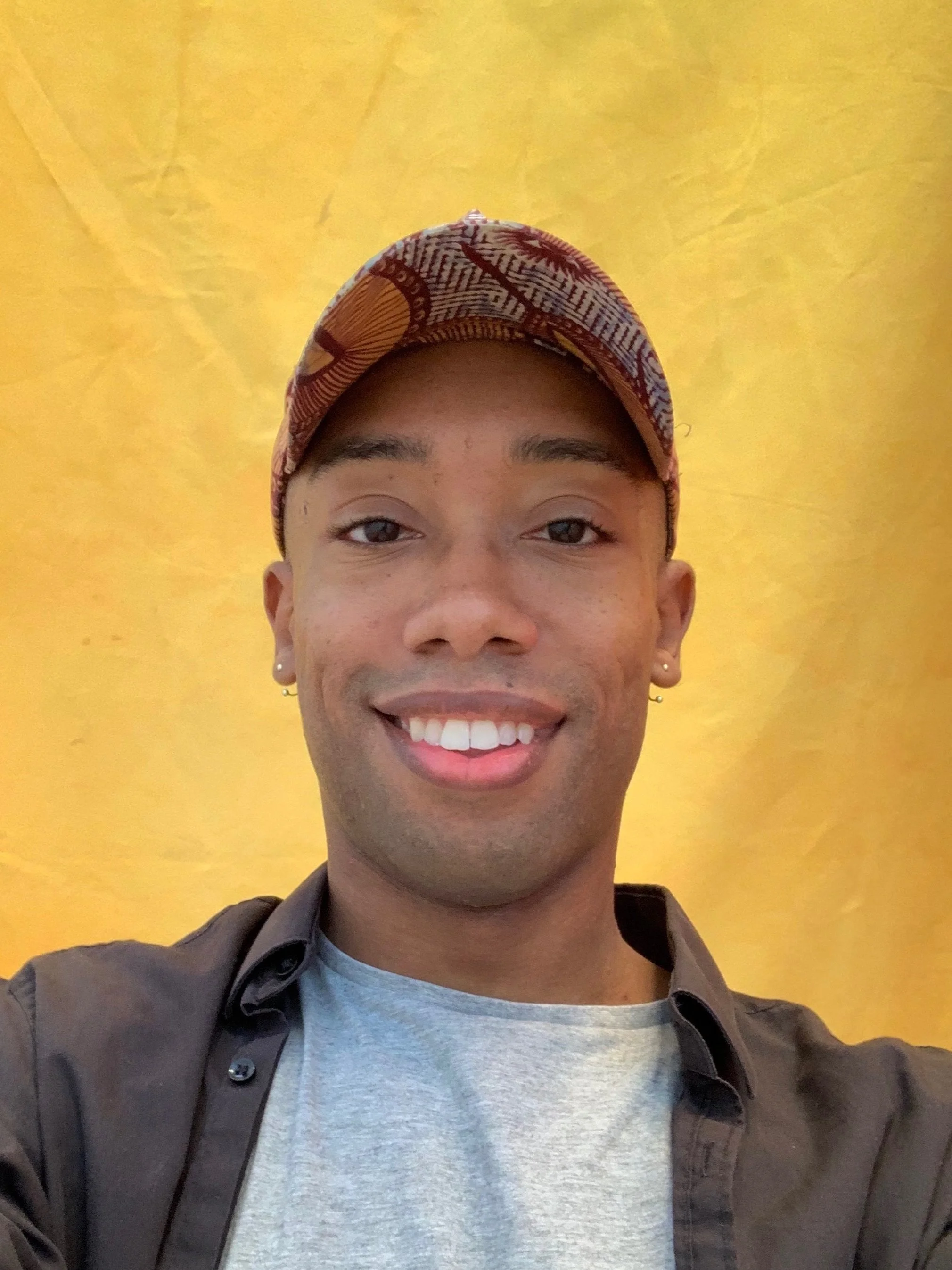 Founder of Pheme AI, Gyasi Patterson, smiling while wearing a patterned cap and black shirt, against a yellow background.