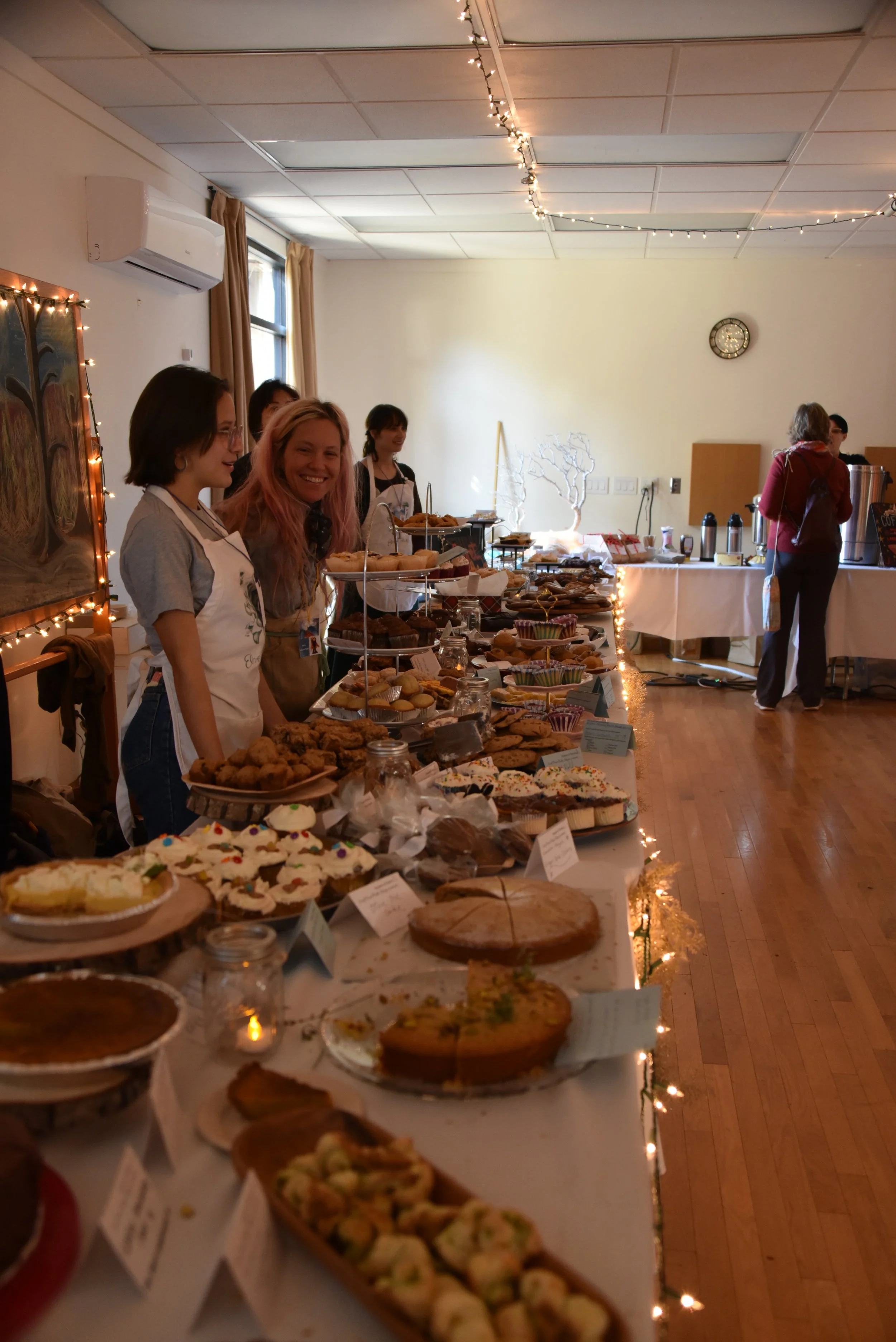 A bake sale with a variety of desserts displayed on long tables. Several people are standing behind the tables, with fairy lights decorating the room. The desserts include cookies, cupcakes, pies, and pastries. The room has a wooden floor and a clock