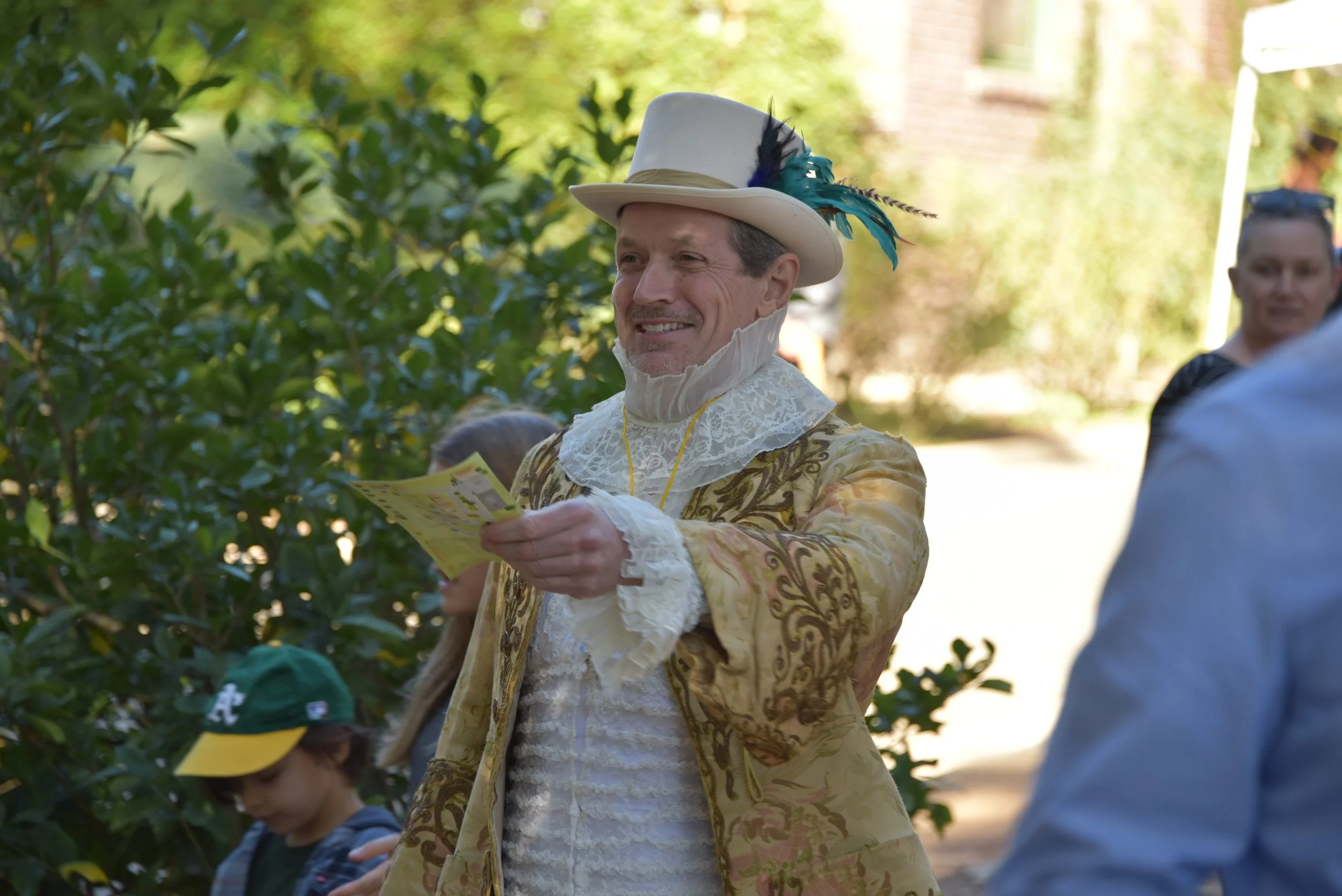 Man in ornate historical costume with feathered hat holding papers, surrounded by greenery.