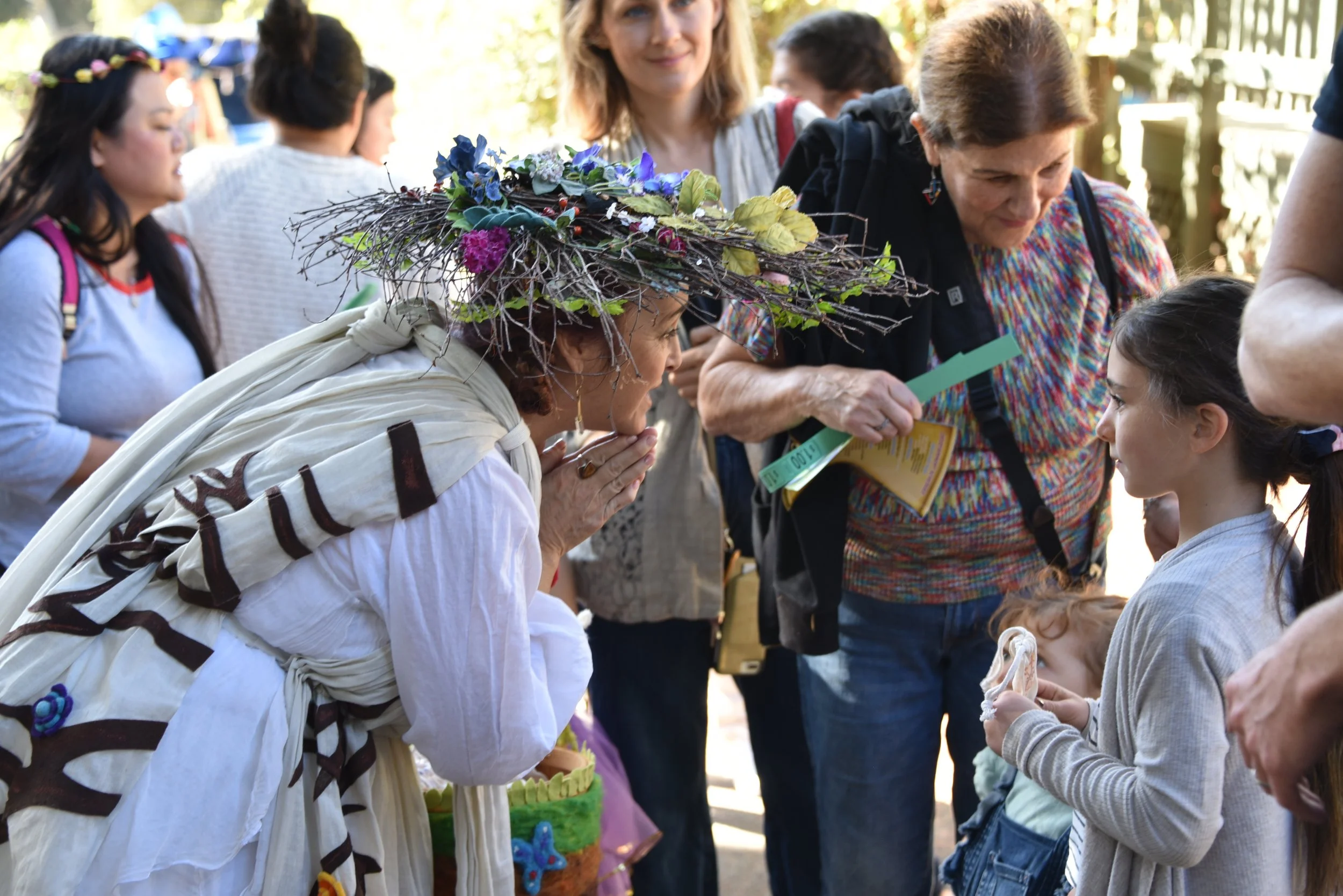 A woman wearing a floral and branch-themed hat is bending down to speak to a child. The child has dark hair and is holding a white item. Several people, including women, are around them interacting in an outdoor setting.