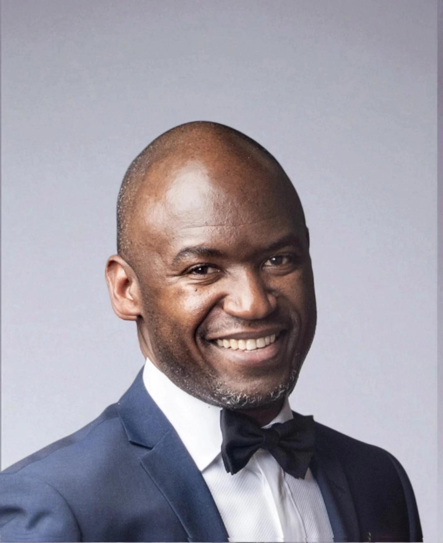 Portrait of a smiling man in formal attire, wearing a dark suit, white shirt, and black bow tie, against a light gray background.