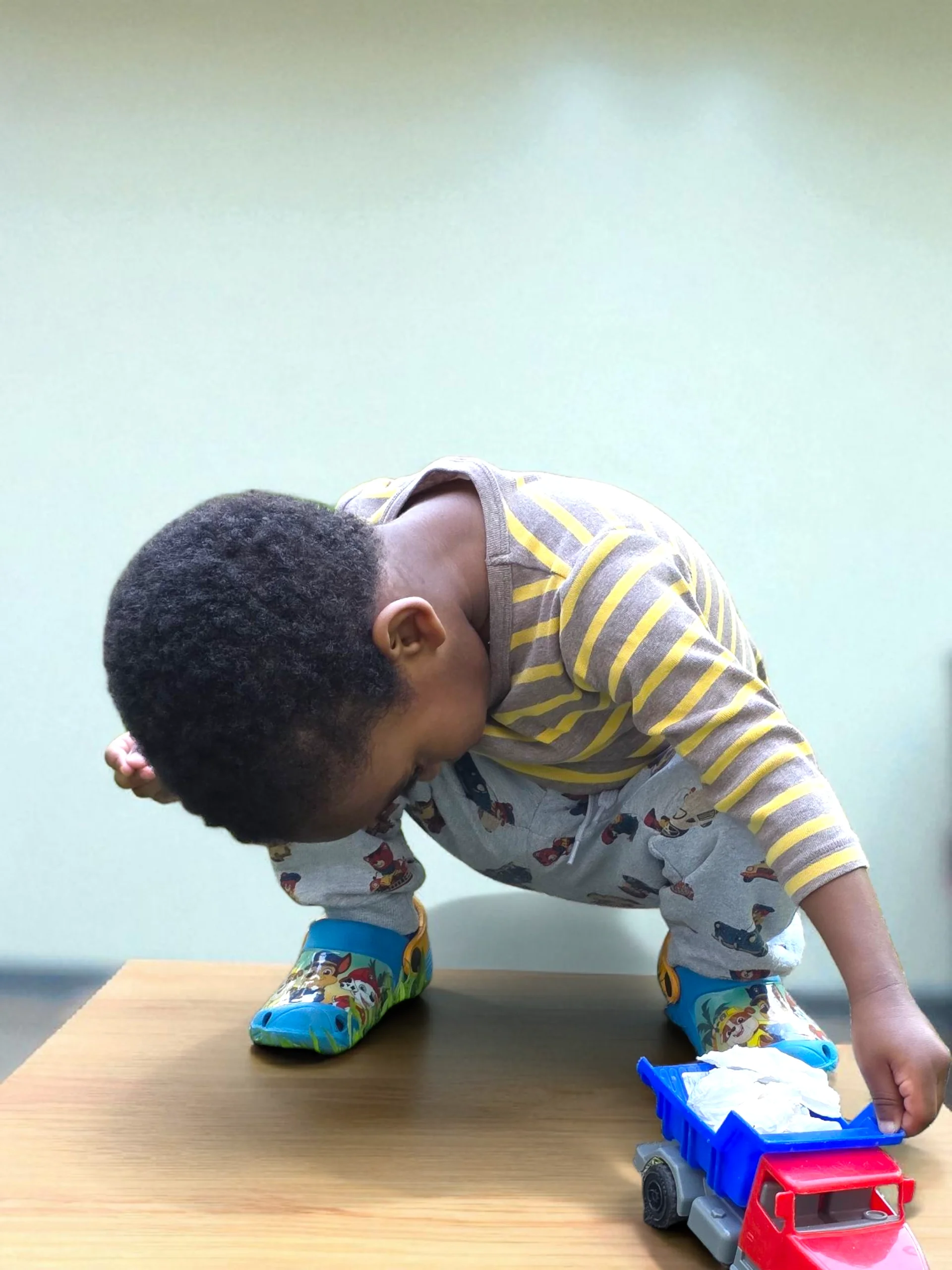 Young boy in pajamas and rain boots on a table, reaching for a toy truck with tissue paper on it.