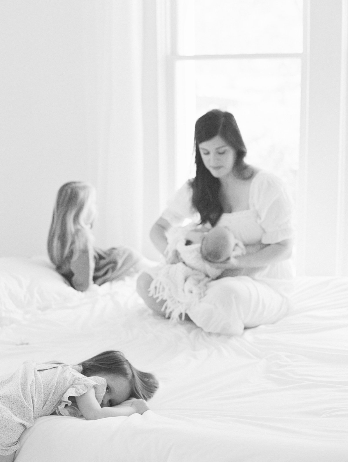 A woman sitting on a bed holding a newborn baby with two young girls nearby, one girl lying on the bed and another sitting and facing the woman, with large windows in the background