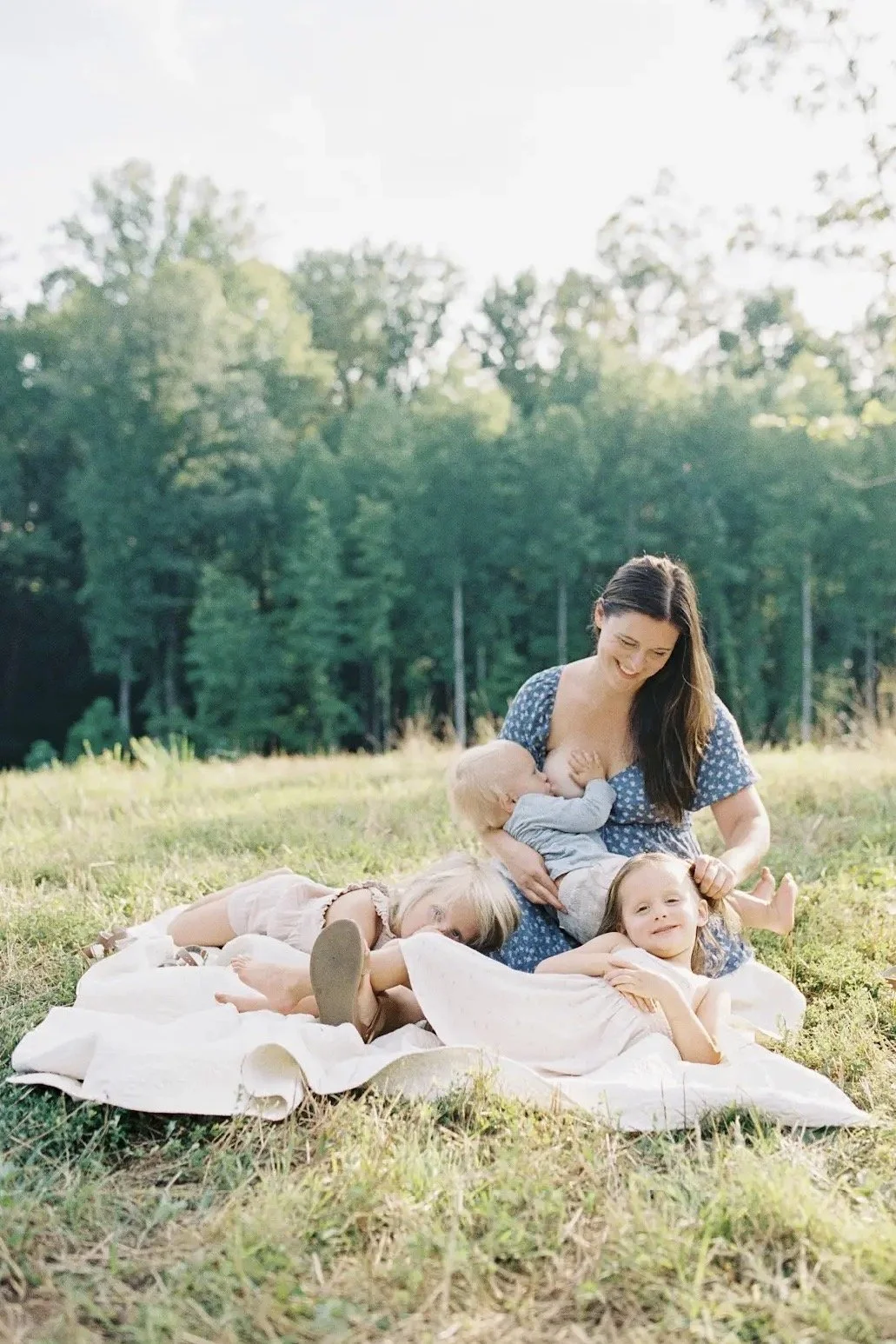 A mother outside on a blanket, breastfeeding a young child while another child rests nearby and a third child looks on with a smile.