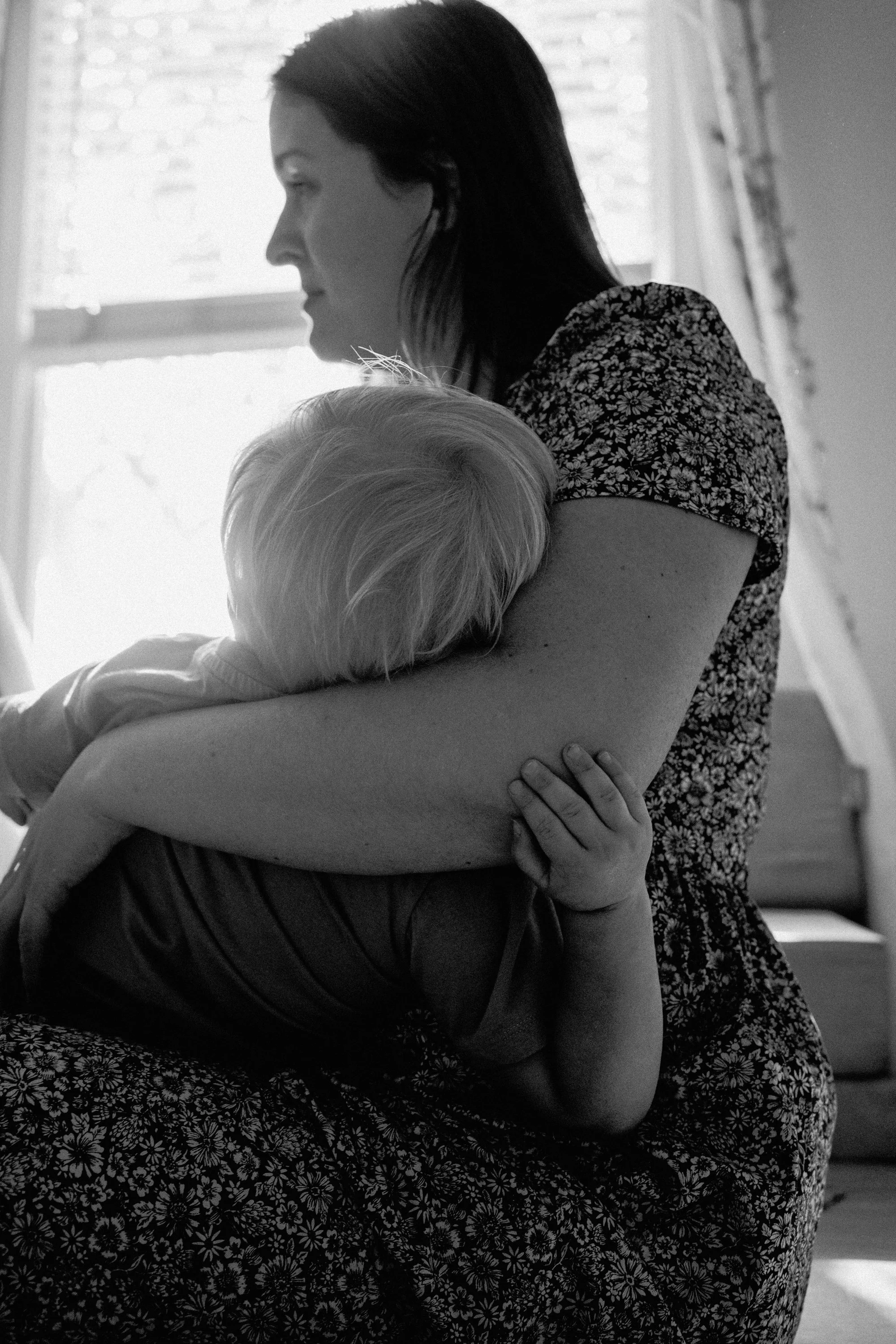 Black and white profile of a mother standing by a window, silhouetted against the light while holding her toddler close in a quiet, reflective moment
