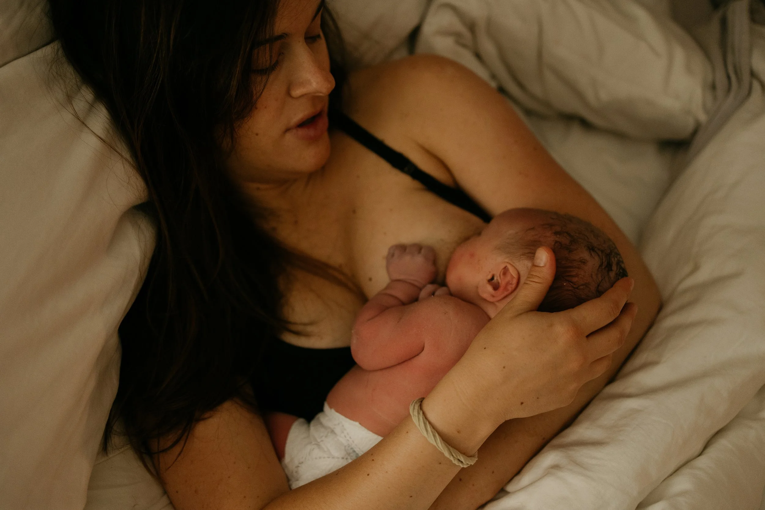 Intimate close-up of a mother lying in bed, holding her newborn baby skin-to-skin against her chest while breastfeeding on white linens.