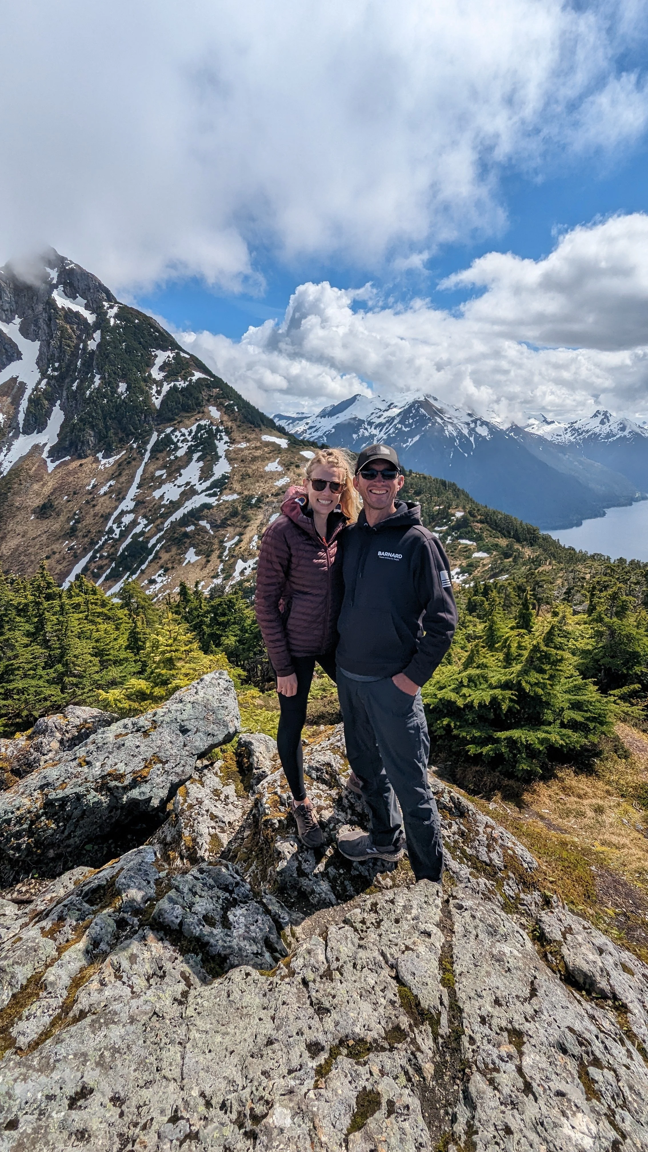 A couple smiling and posing on a rocky mountain trail, with snow-capped peaks, a lake, and lush green trees in the background on a partly cloudy day.