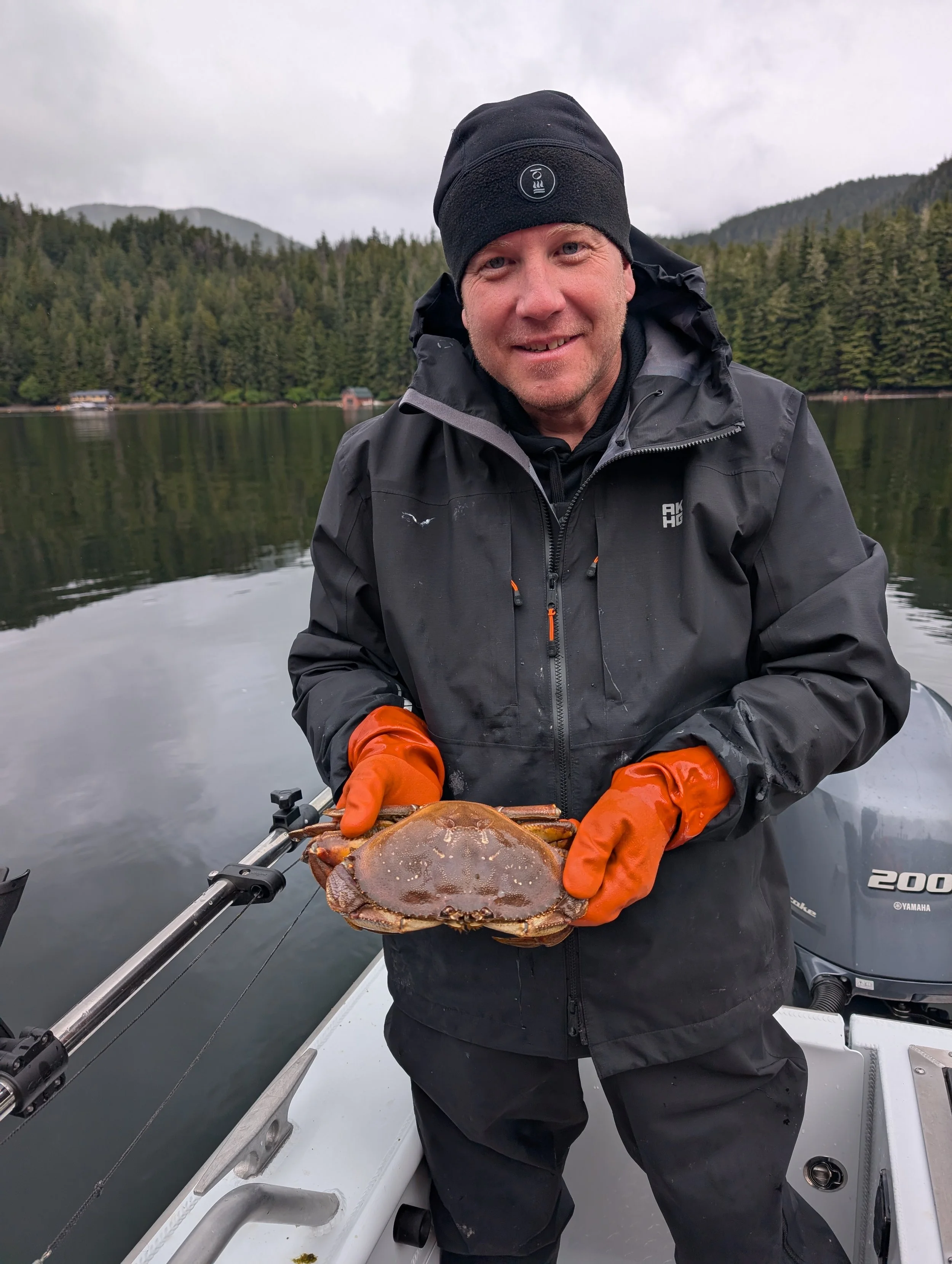 A man in a black jacket and beanie holding a large crab while on a boat in a lake with a forested shoreline and mountains in the background.
