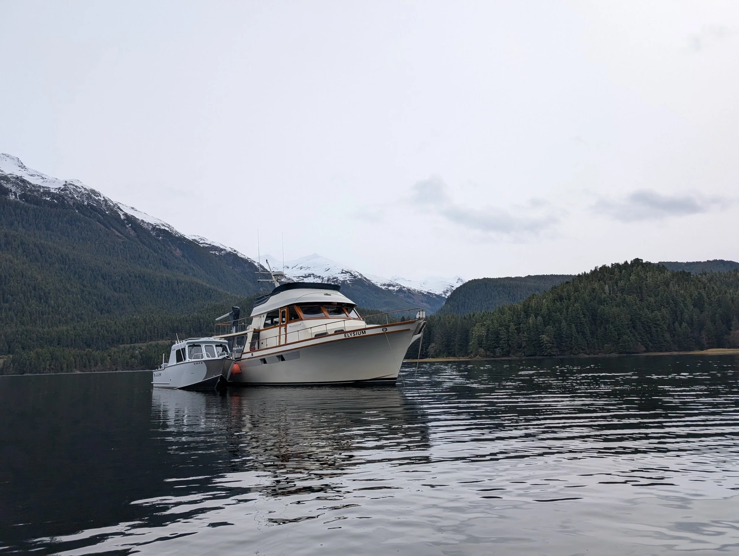 Boats anchored on a calm lake with snow-capped mountains and pine trees in the background under a cloudy sky.