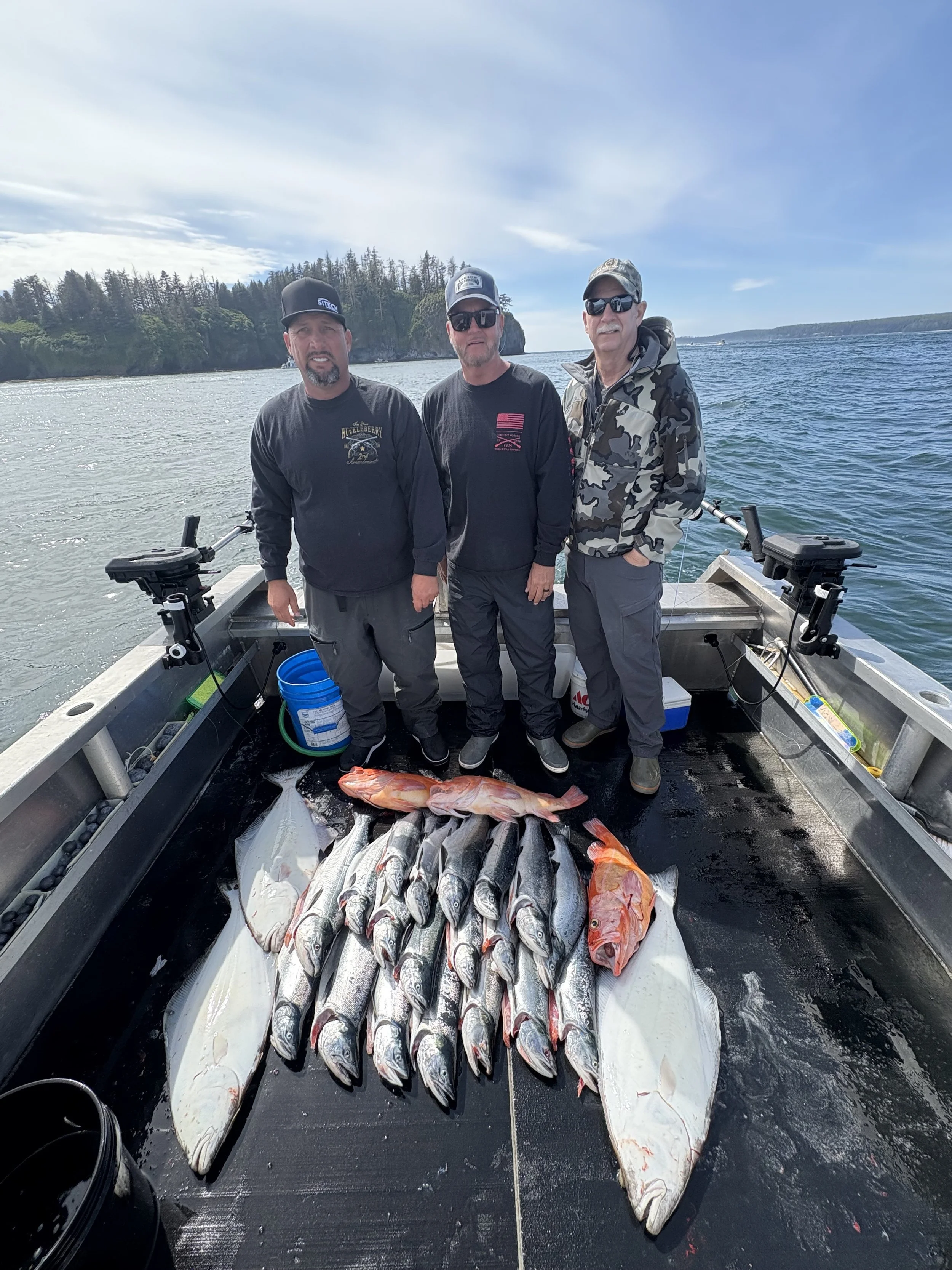 Three men standing on a boat with a catch of fish laid out on the floor, including several large fish and some smaller fish, with water and a forested shoreline in the background.