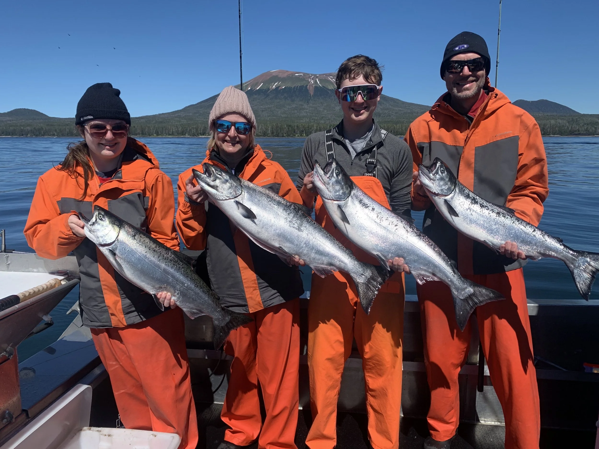 Five people standing on a boat holding large fish, with a mountain and lake in the background.