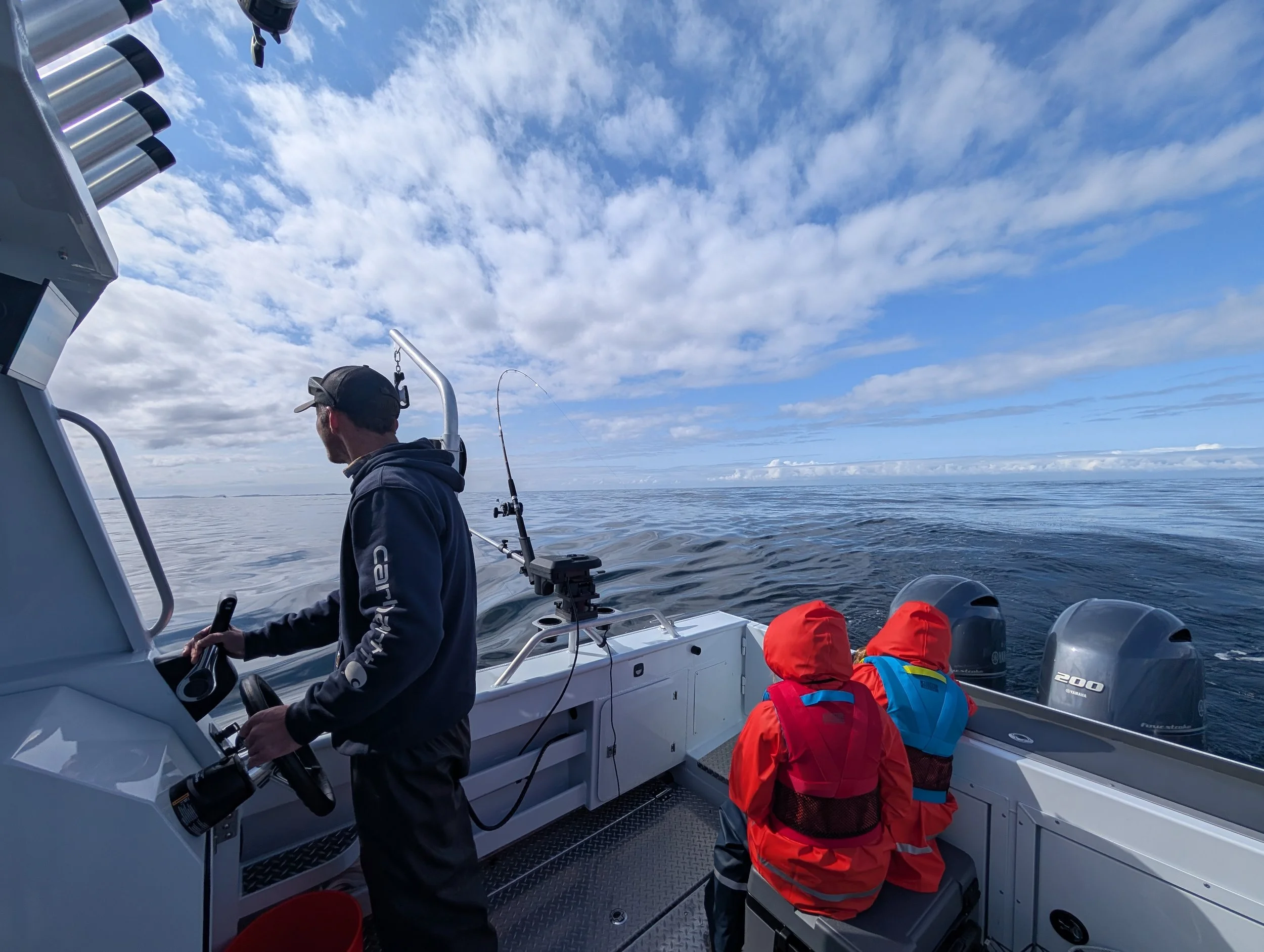 A man steering a boat with two children on board, one in a red jacket and blue life vest, on a large body of water under a partly cloudy sky.