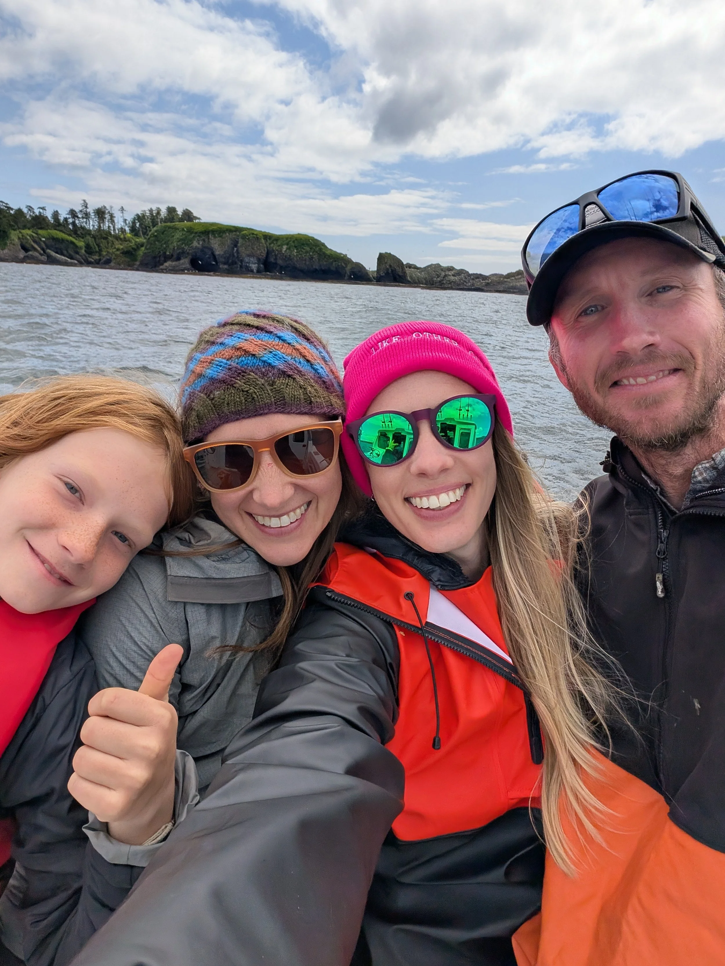 A group of four people smiling and taking a selfie outdoors on a cloudy day with rocky cliffs and water in the background.