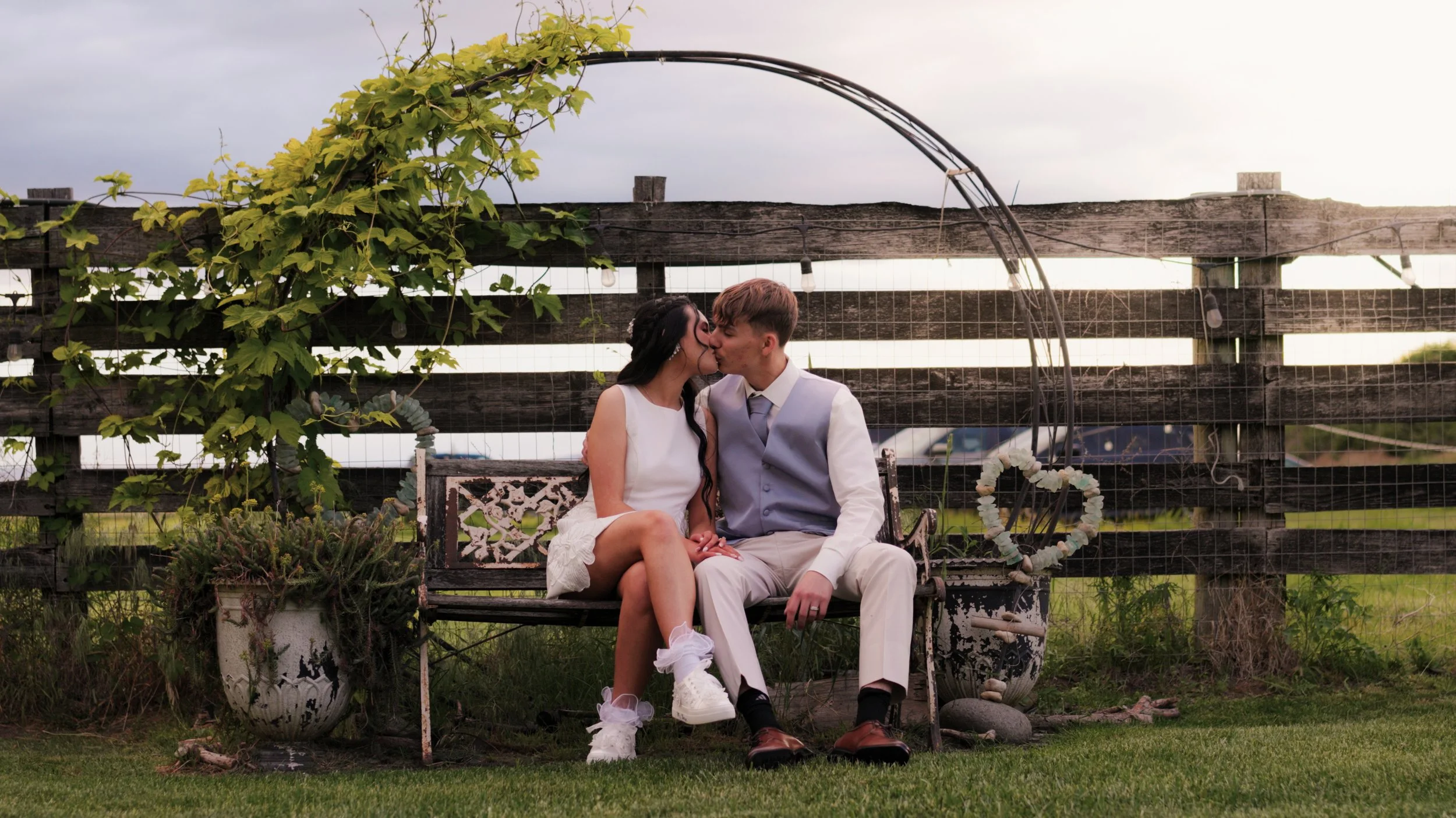 A couple sitting on a bench, sharing a kiss, outdoors in front of a wooden fence decorated with green climbing plants, with potted plants and a heart-shaped wreath nearby.