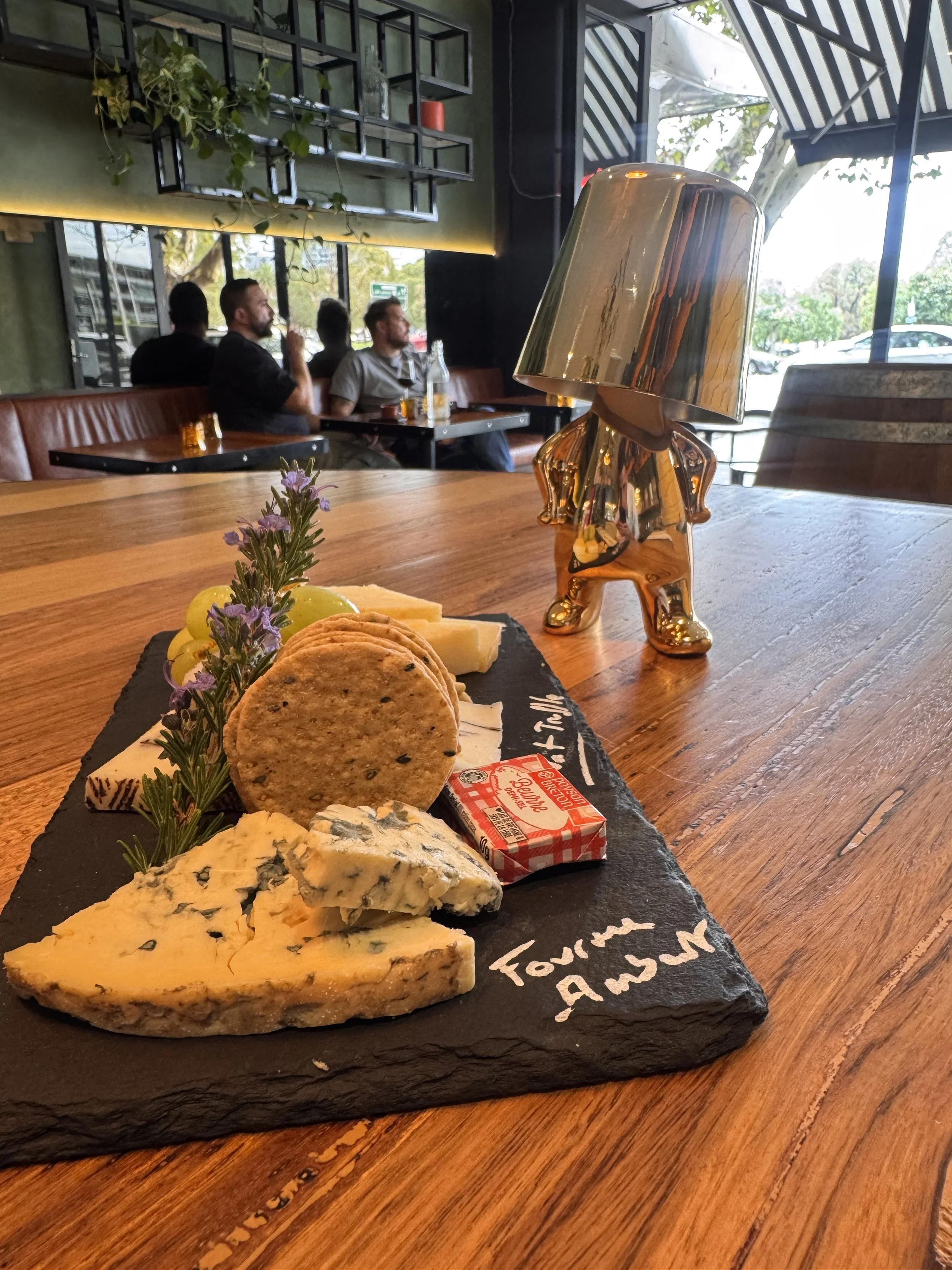 A cheese and crackers platter with herbs and butter on a black slate plate, a metallic table lamp, and a woman figurine in a restaurant.