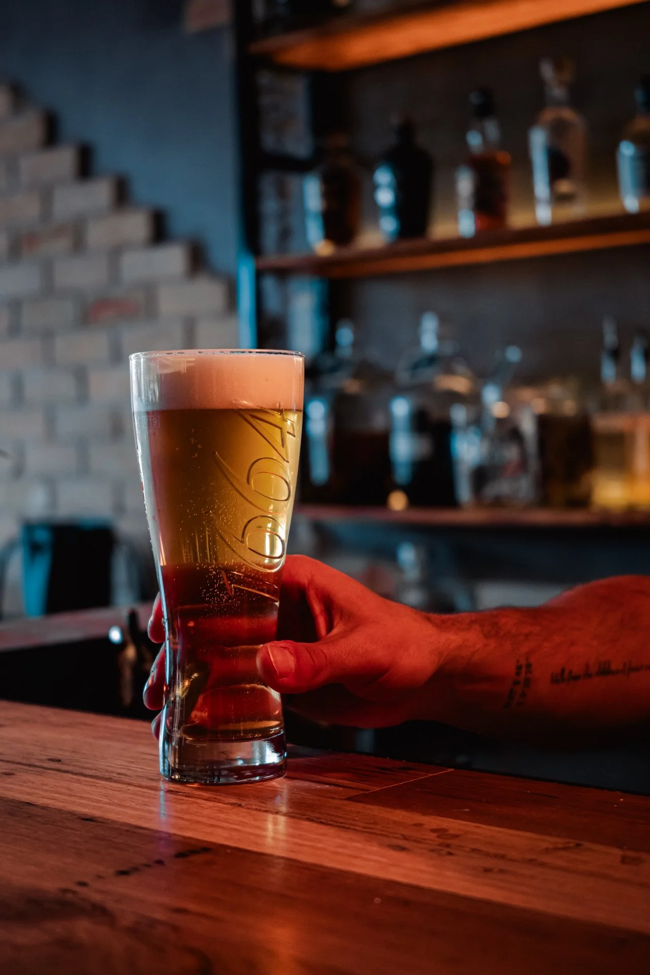 A person with a tattooed arm holding a glass of beer at a bar or pub, with shelves of bottles in the background.