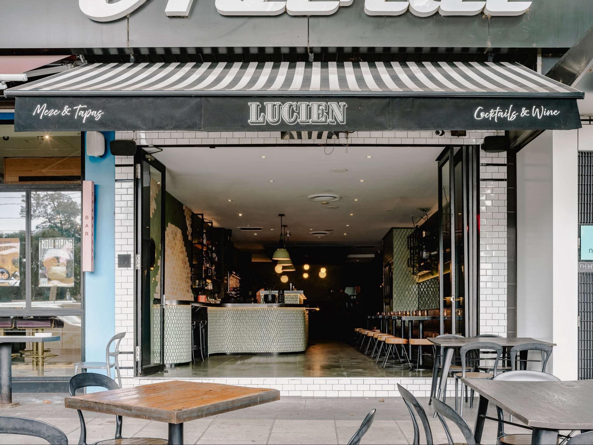 Outdoor view of Lucien bar and restaurant, featuring a striped awning, open seating area, and modern interior design.