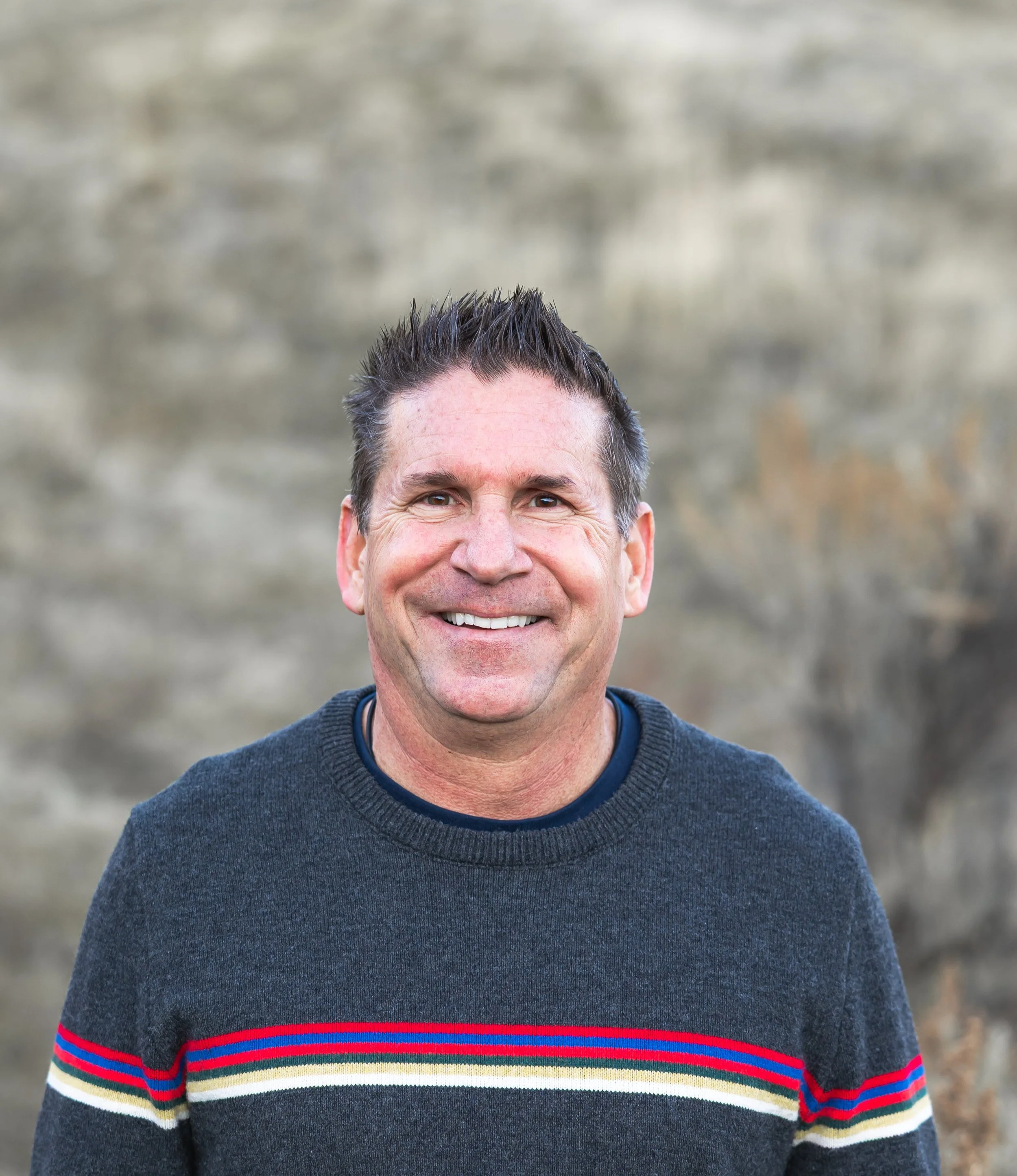 A smiling middle-aged man with short dark hair, wearing a dark sweater with colorful stripes, standing outdoors in front of a blurred stone wall background.