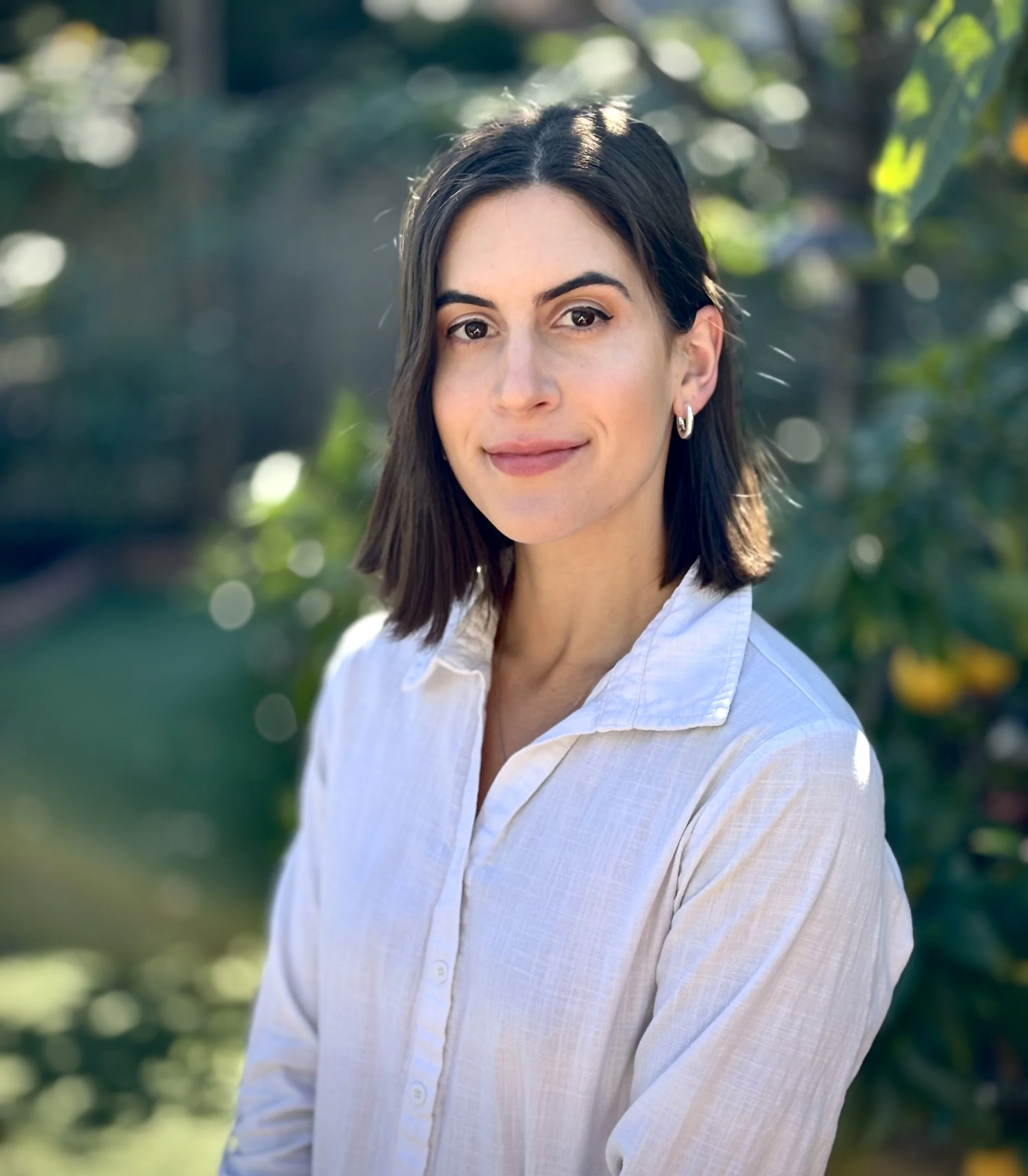 A woman with shoulder-length dark brown hair, wearing a white shirt and hoop earrings, standing outdoors in a garden with green plants and sunlight in the background.