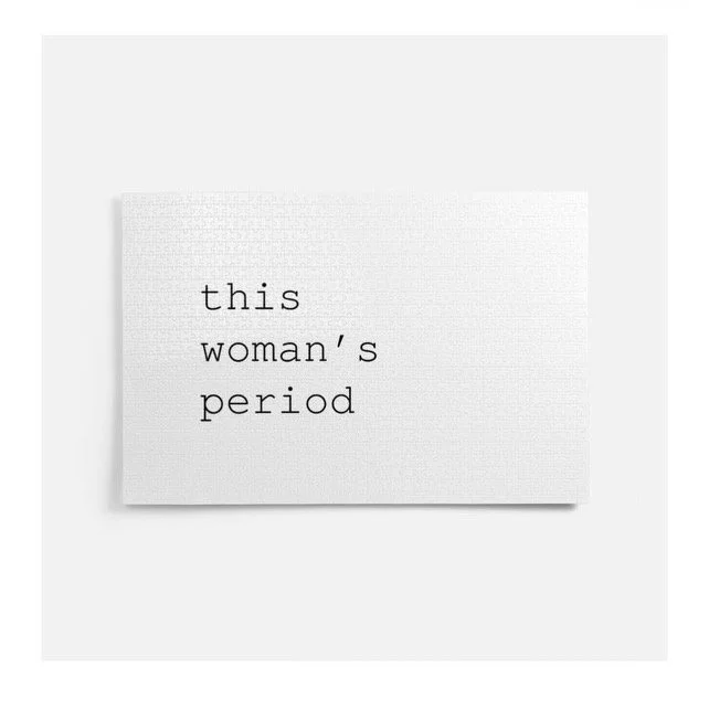 A white card with the text 'this woman's period' typed in black ink, placed on a flat surface.