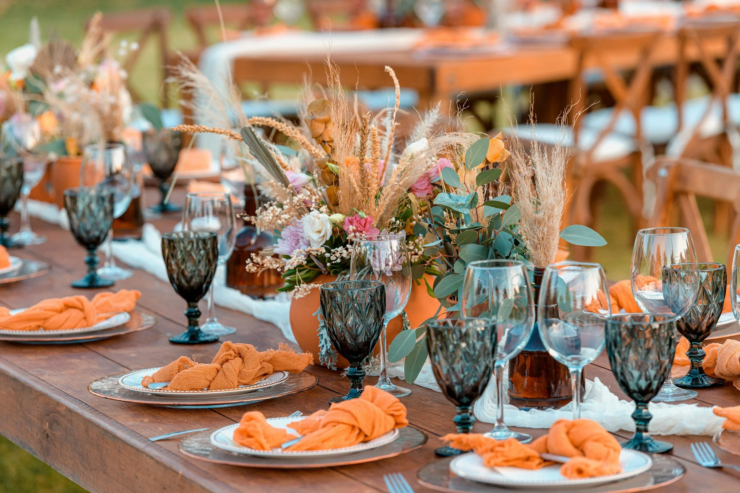 A table set for an outdoor event decorated with orange napkins, glassware, and a floral centerpiece featuring pink, white, and green foliage and dried grasses.