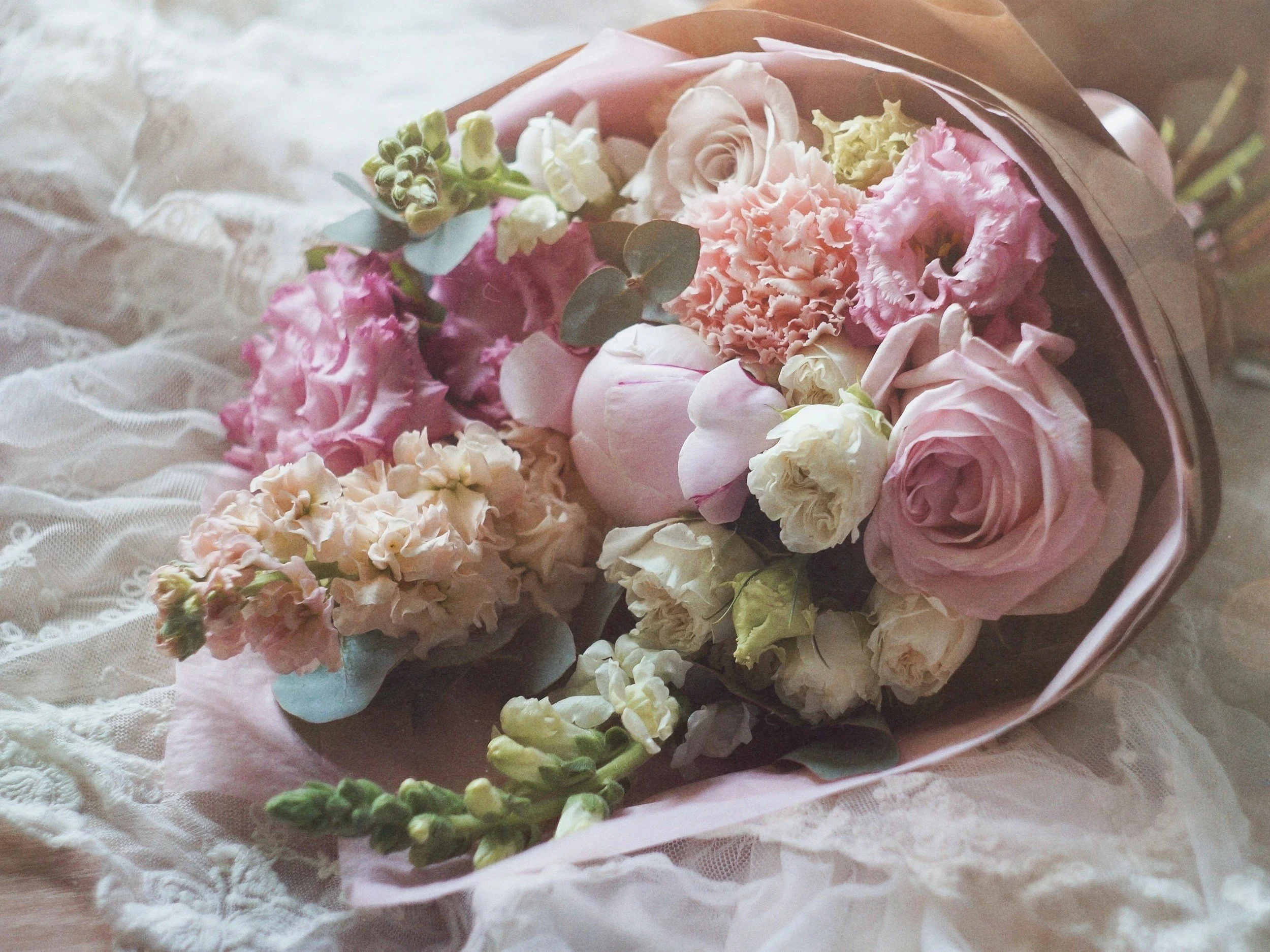 A bouquet of pink and white flowers wrapped in pink paper resting on lace fabric.