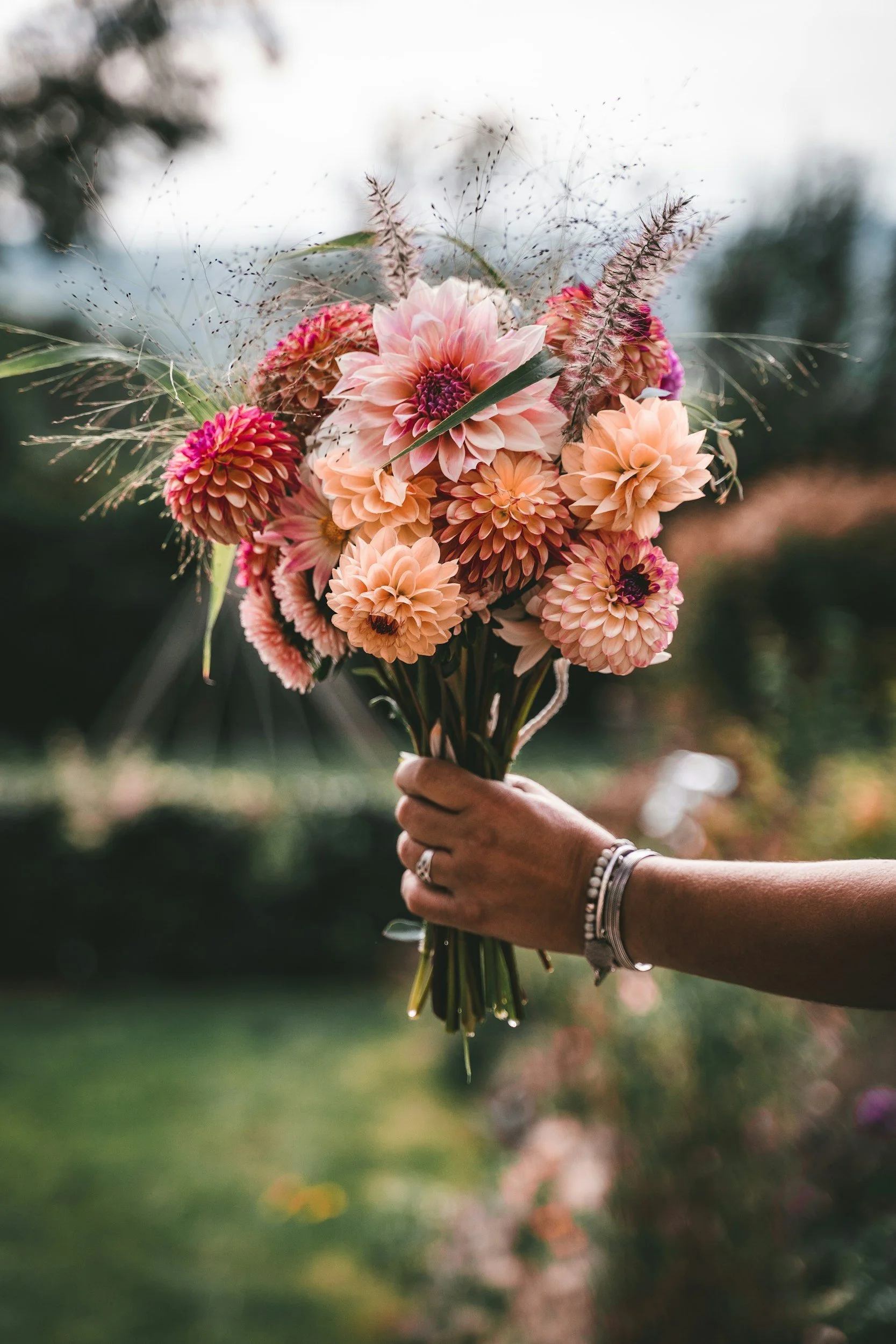Person holding a bouquet of pink, peach, and purple flowers with greenery and decorative feathers outside.