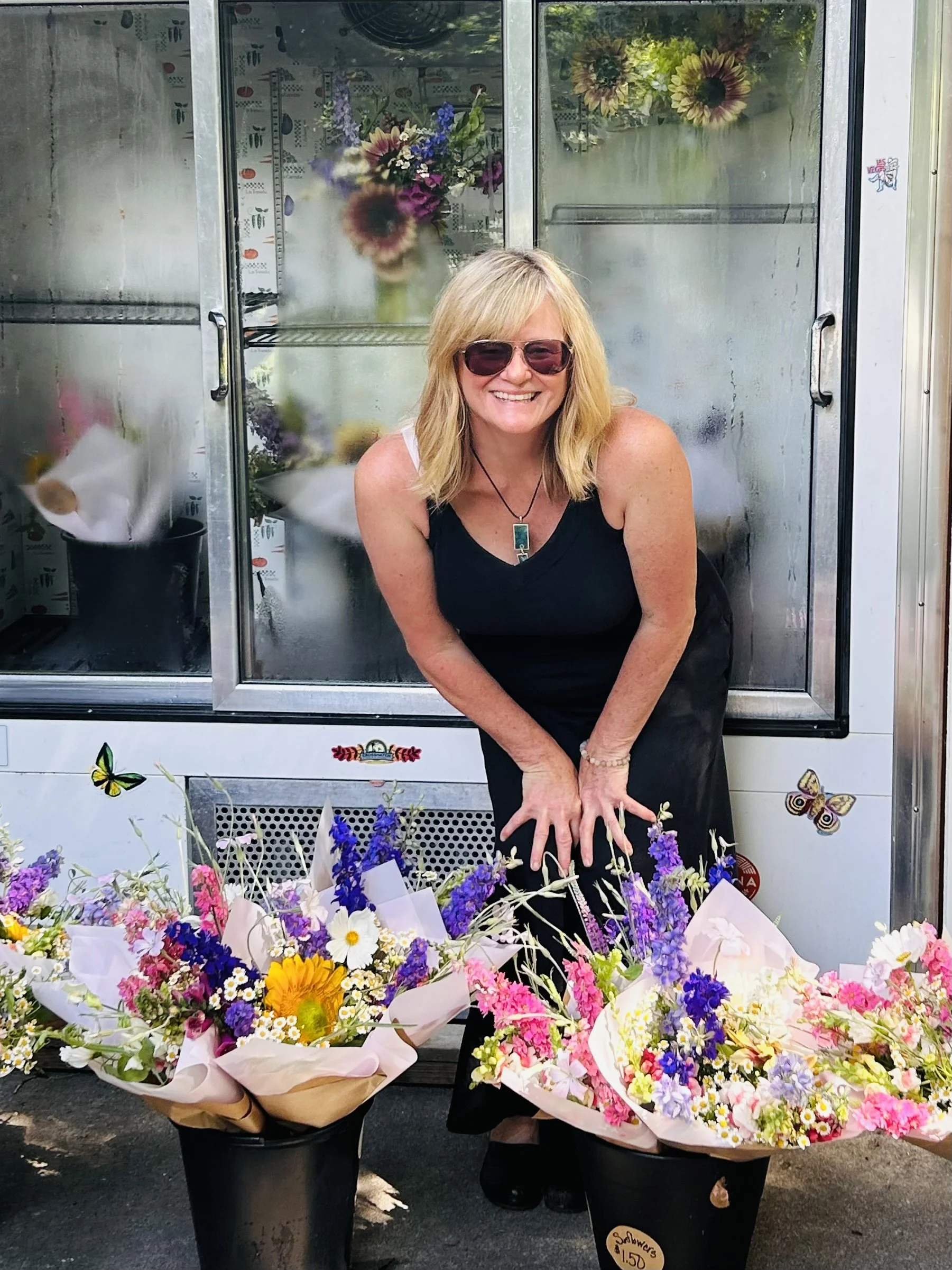 A woman with blonde hair wearing sunglasses and a black sleeveless top, smiling, is surrounded by four buckets of colorful flowers, and standing in front of a glass flower fridge.