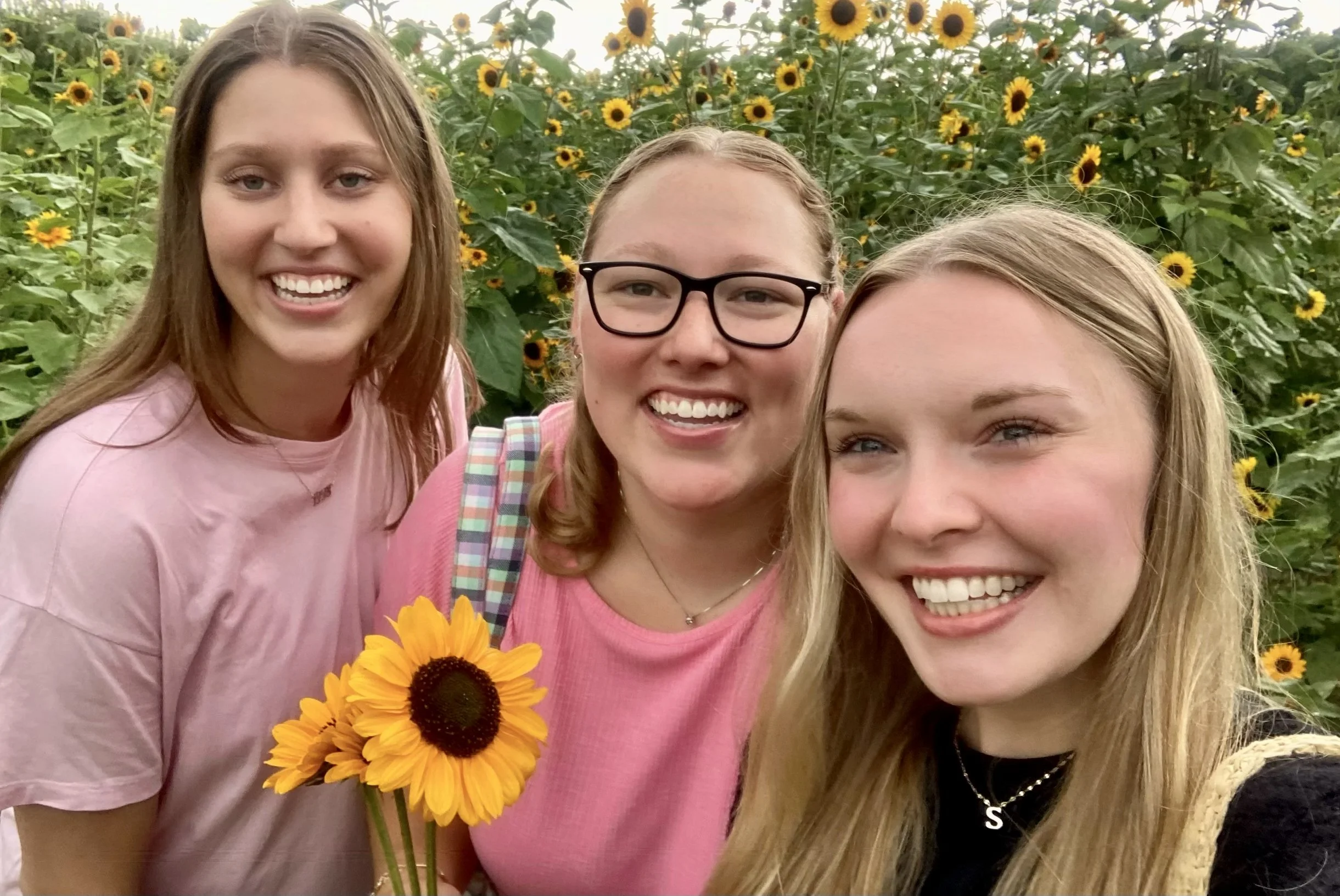 Three young women smiling in a sunflower field, one holding a sunflower.