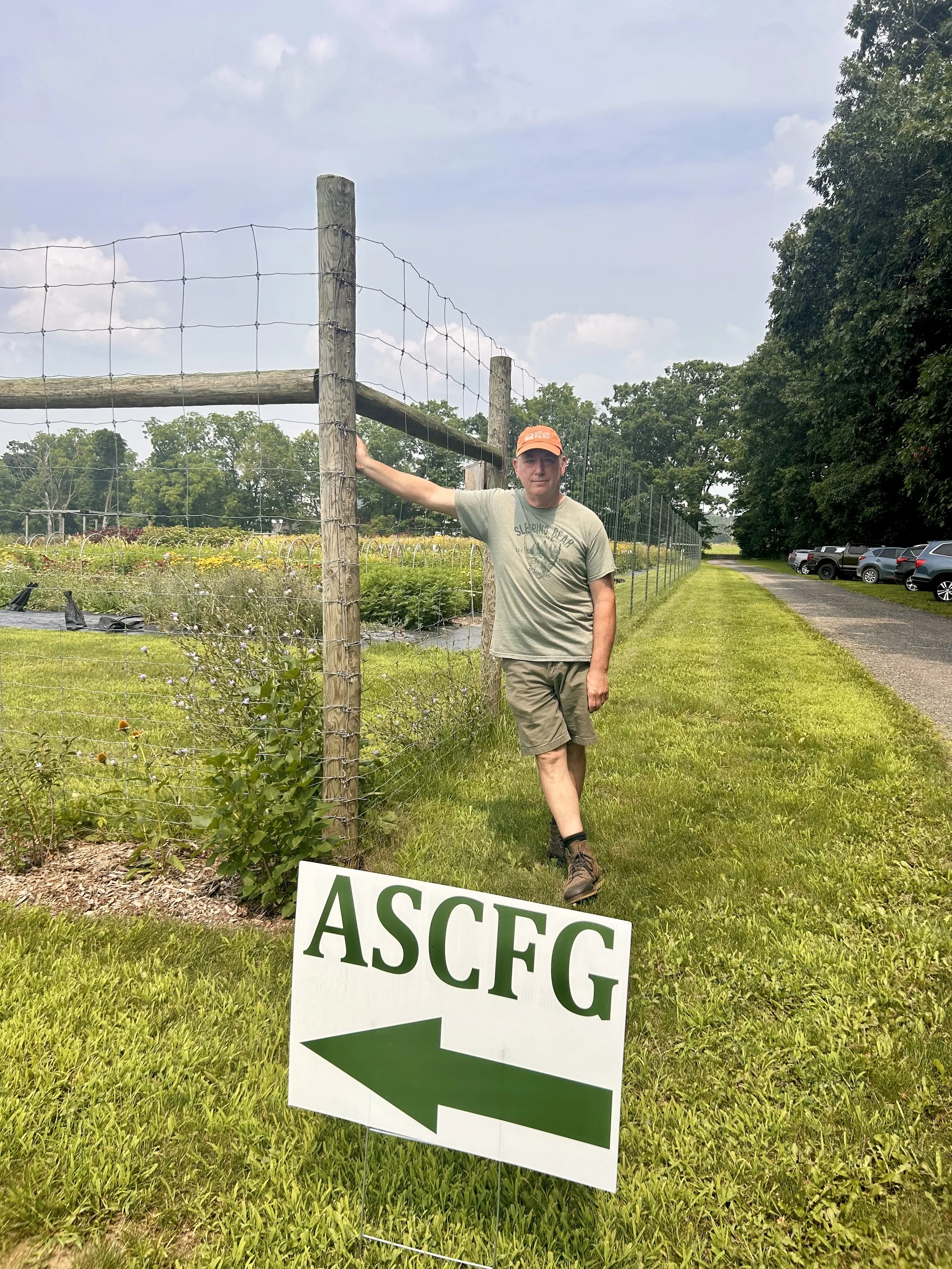 A man wearing a tan t-shirt, khaki shorts, and a bright orange cap stands outdoors next to a wire fence and a sign with the text 'ASCFG' and a left-pointing arrow. The setting appears to be a rural area with parking on the right and a vegetable garden beyond the fence.