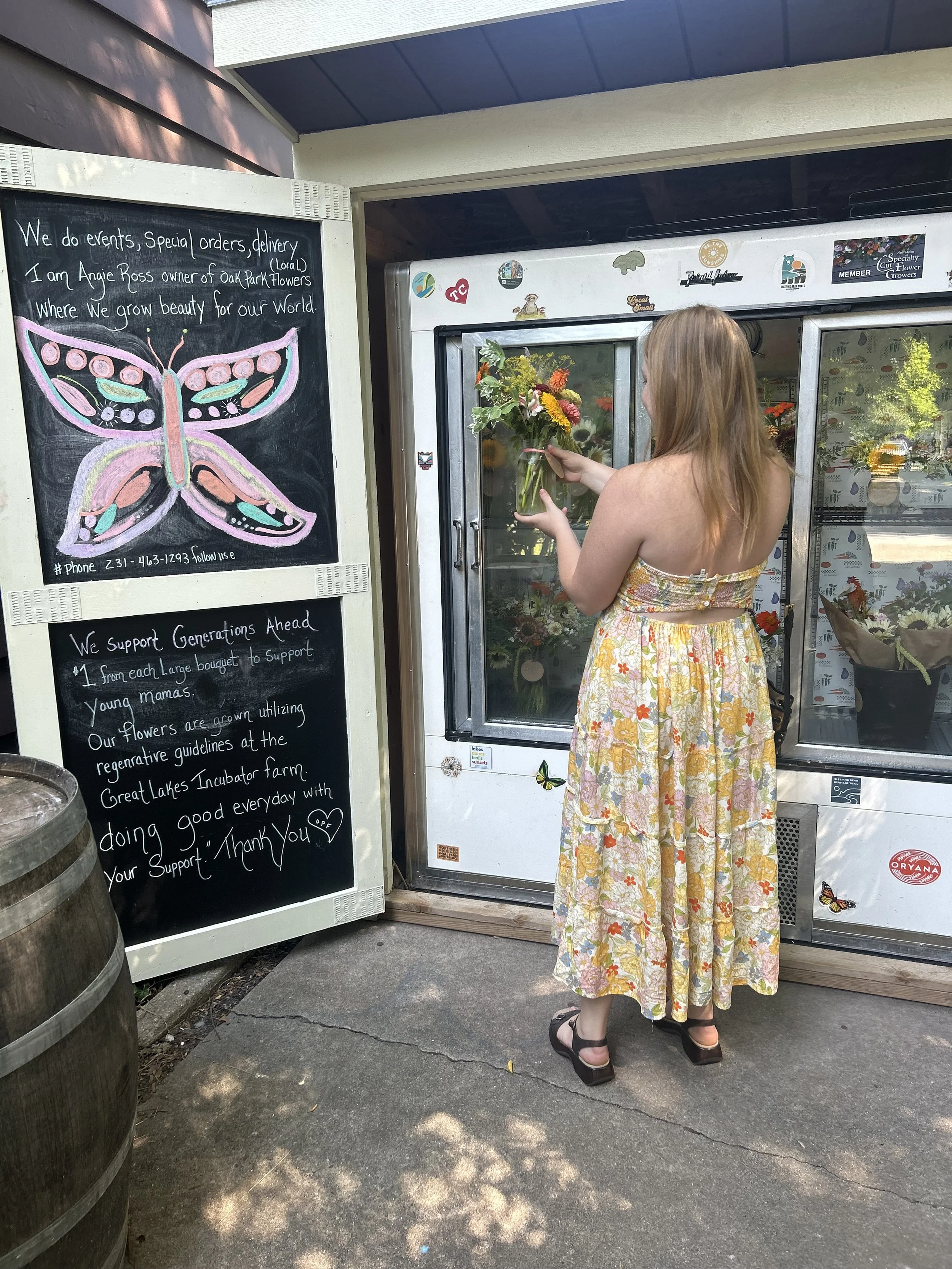 A woman wearing a floral dress and black sandals placing a bouquet of flowers inside a cooler outside a flower shop.