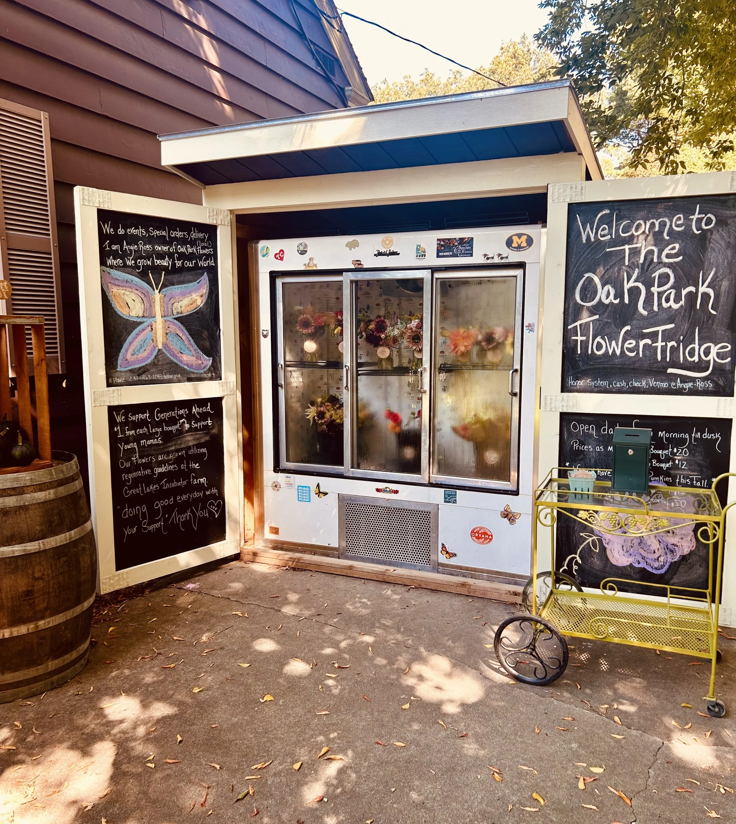 Small flower refrigerator with two glass doors, decorated with stickers, and flowers inside. Black chalkboard signs on both sides with handwritten messages and drawings, including a butterfly illustration on the left, welcoming message on the right, and details about flower sales and support for the community.