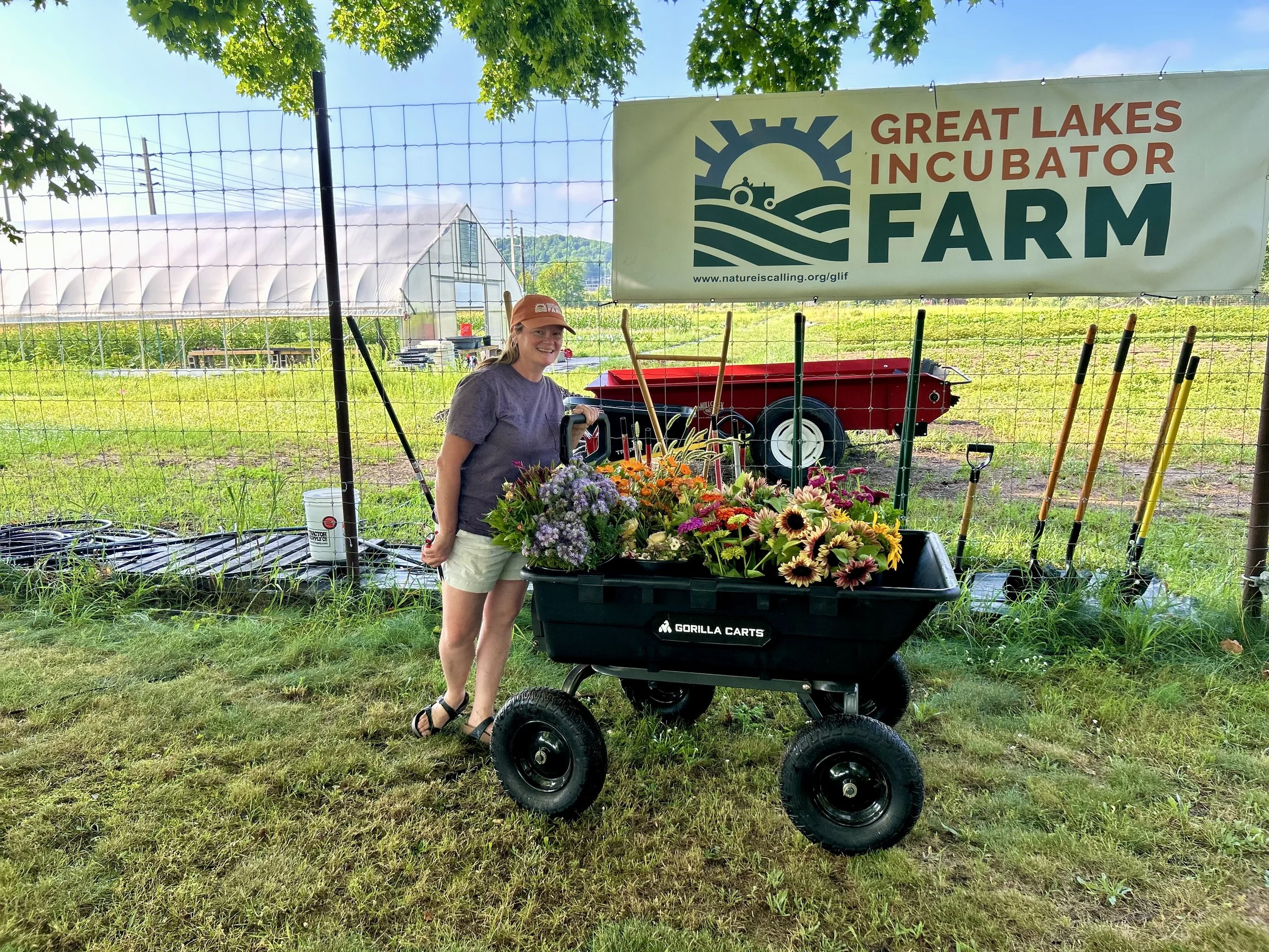 A woman standing next to a wagon filled with colorful flowers at Great Lakes Incubator Farm, with gardening tools and a garden in the background.